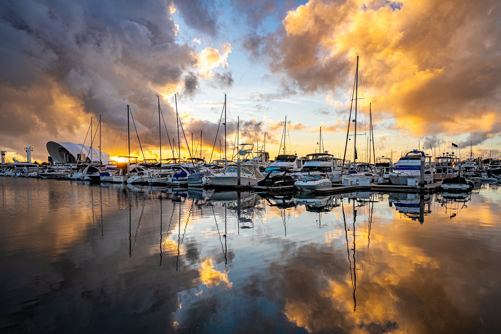 Boats docked at a marina with a colorful sky and clouds reflected in calm water.