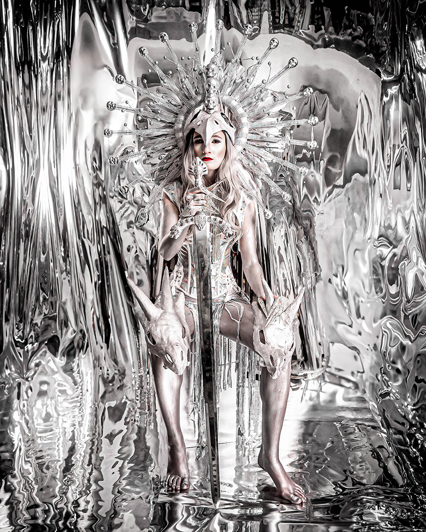 Person in elaborate silver costume with headpiece, sitting in a reflective metallic environment, holding a glass, with long hair and makeup.
