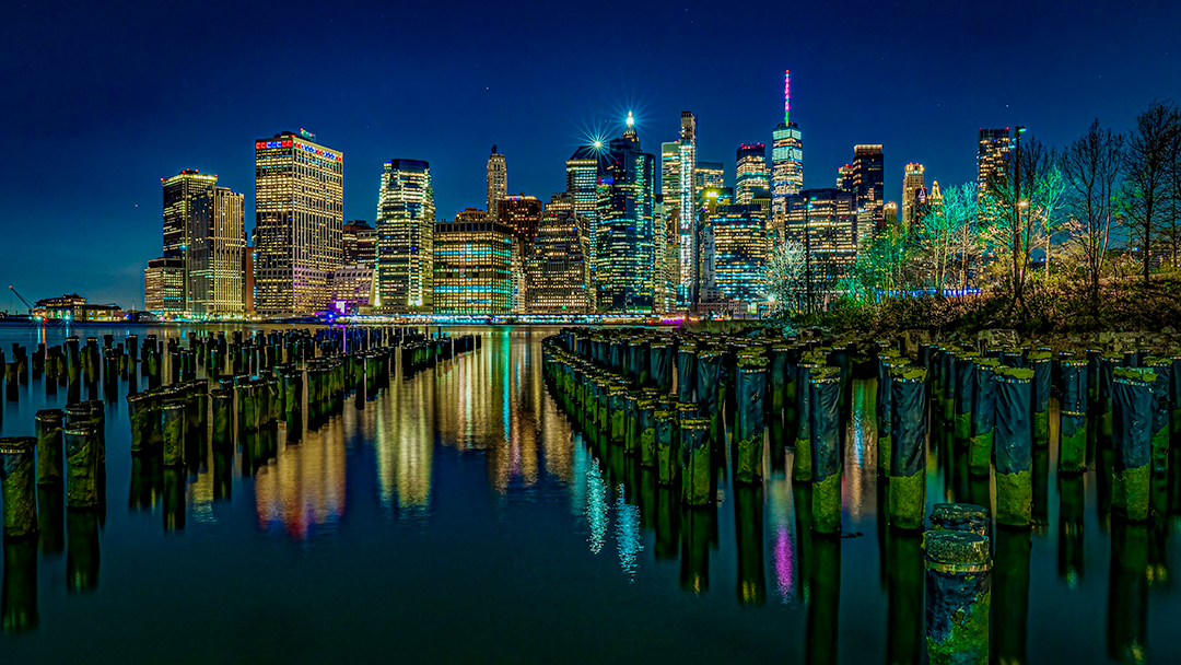 Night city skyline with illuminated buildings reflected in water, wooden posts in foreground, trees on right side.