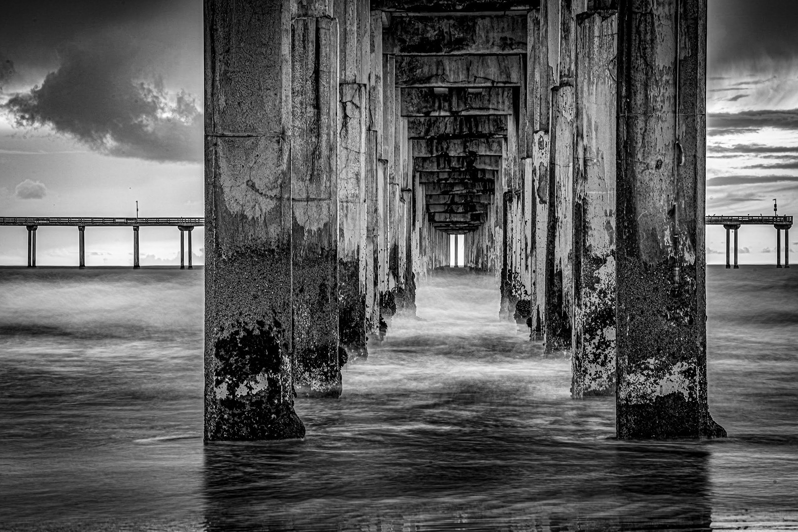 View underneath a pier with concrete pillars extending into the water, leading to a distant horizon, in black and white.