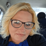 Woman with blonde hair and glasses sitting in a car, smiling, wearing a blue top and black jacket.