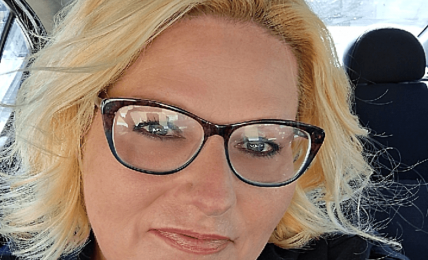 Woman with blonde hair and glasses sitting in a car, smiling, wearing a blue top and black jacket.