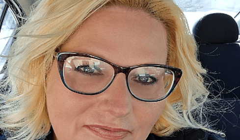 Woman with blonde hair and glasses sitting in a car, smiling, wearing a blue top and black jacket.