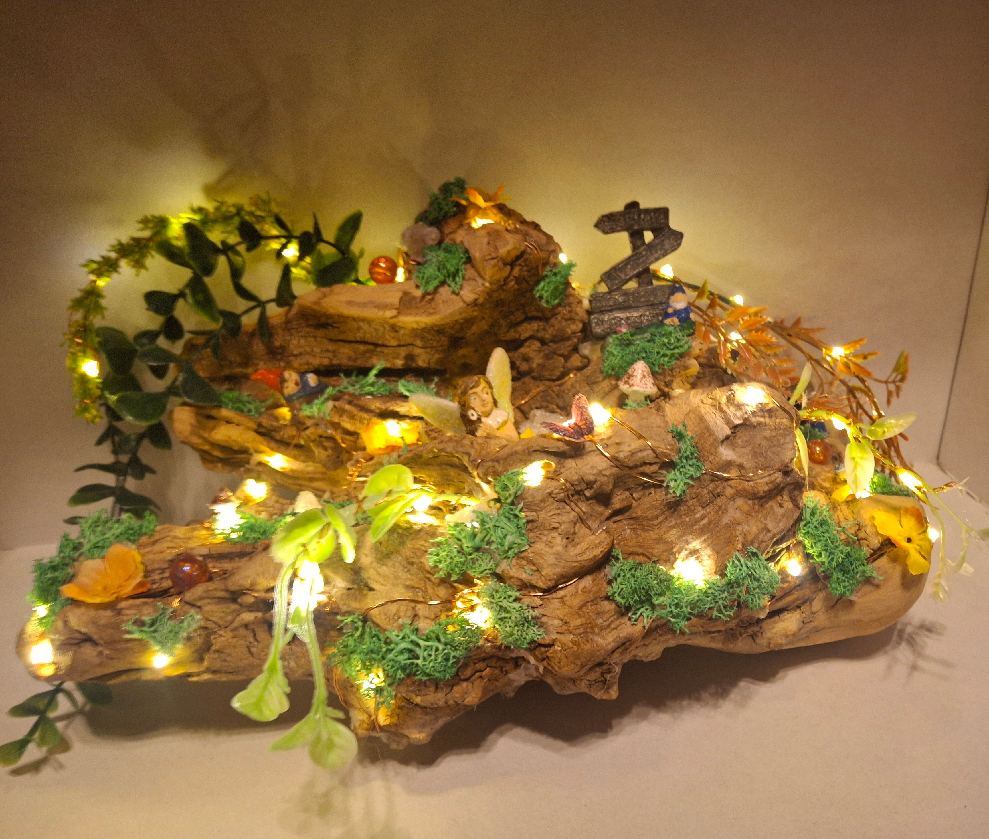 Decorative arrangement with rocks, greenery, and string lights, featuring small figurines and ornaments, on a neutral background.