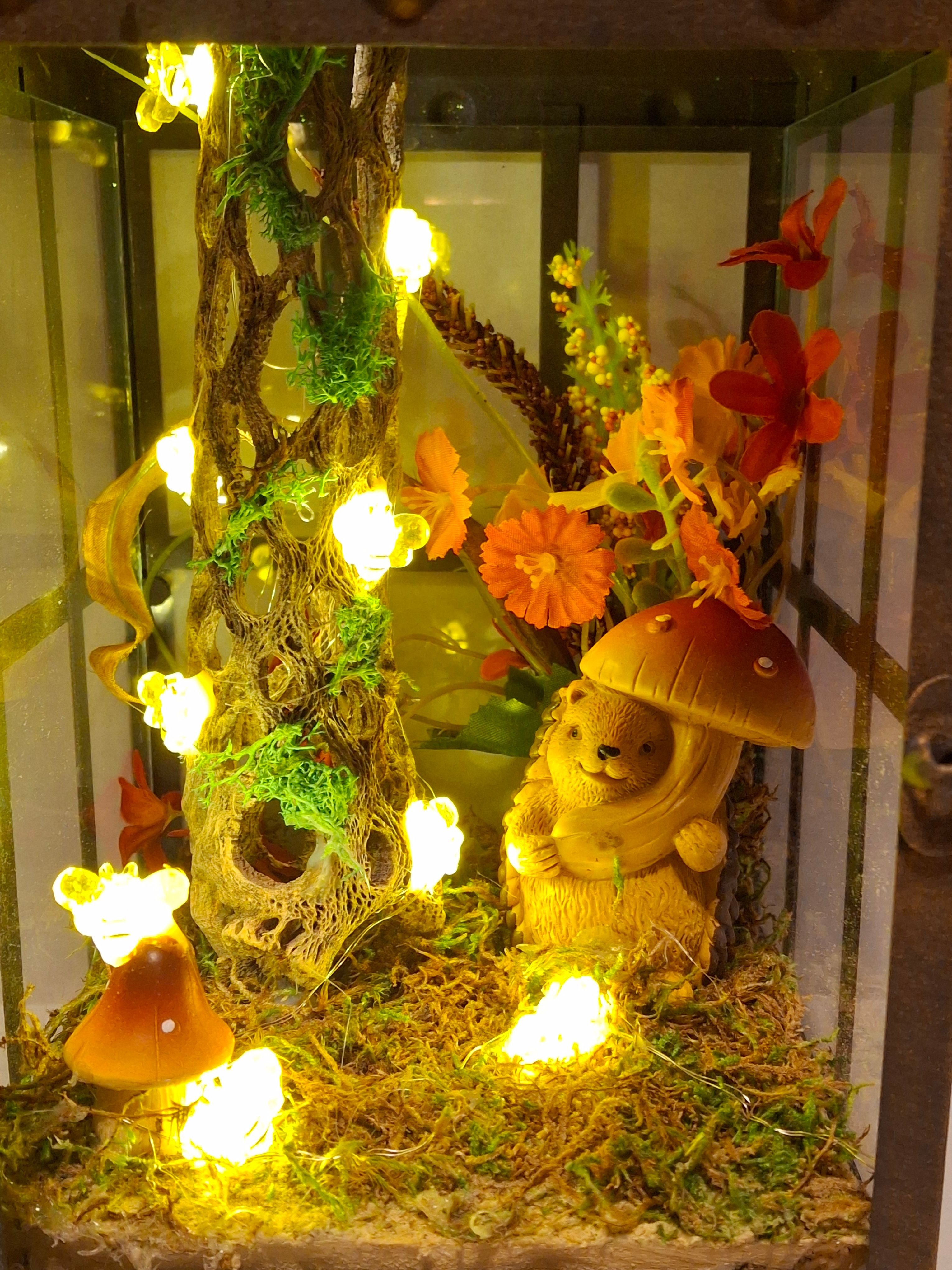 Decorative arrangement with flowers, mushrooms, and a small animal figure, illuminated by warm yellow lights, on a glass surface.
