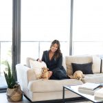 Woman sitting on a white sofa with two dogs, in a bright room with large windows and a potted plant.