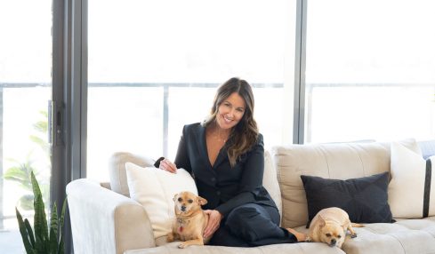 Woman sitting on a white sofa with two dogs, in a bright room with large windows and a potted plant.