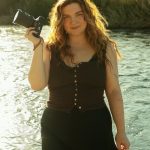 Young woman with curly hair holding a camera, standing near water with a grassy background.