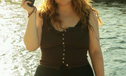 Young woman with curly hair holding a camera, standing near water with a grassy background.