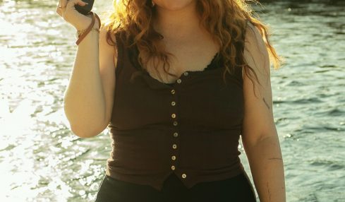 Young woman with curly hair holding a camera, standing near water with a grassy background.
