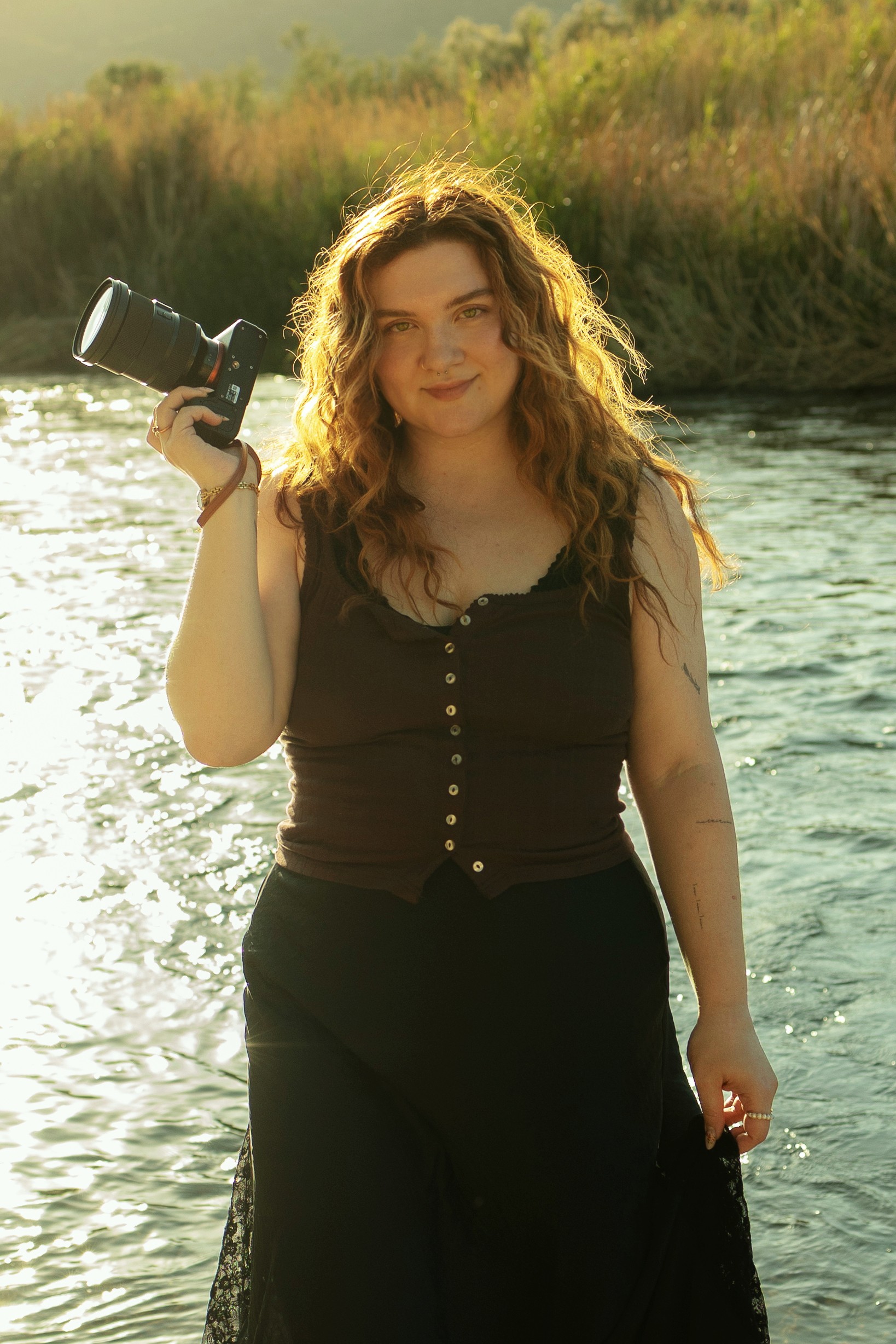 Young woman with curly hair holding a camera, standing near water with a grassy background.