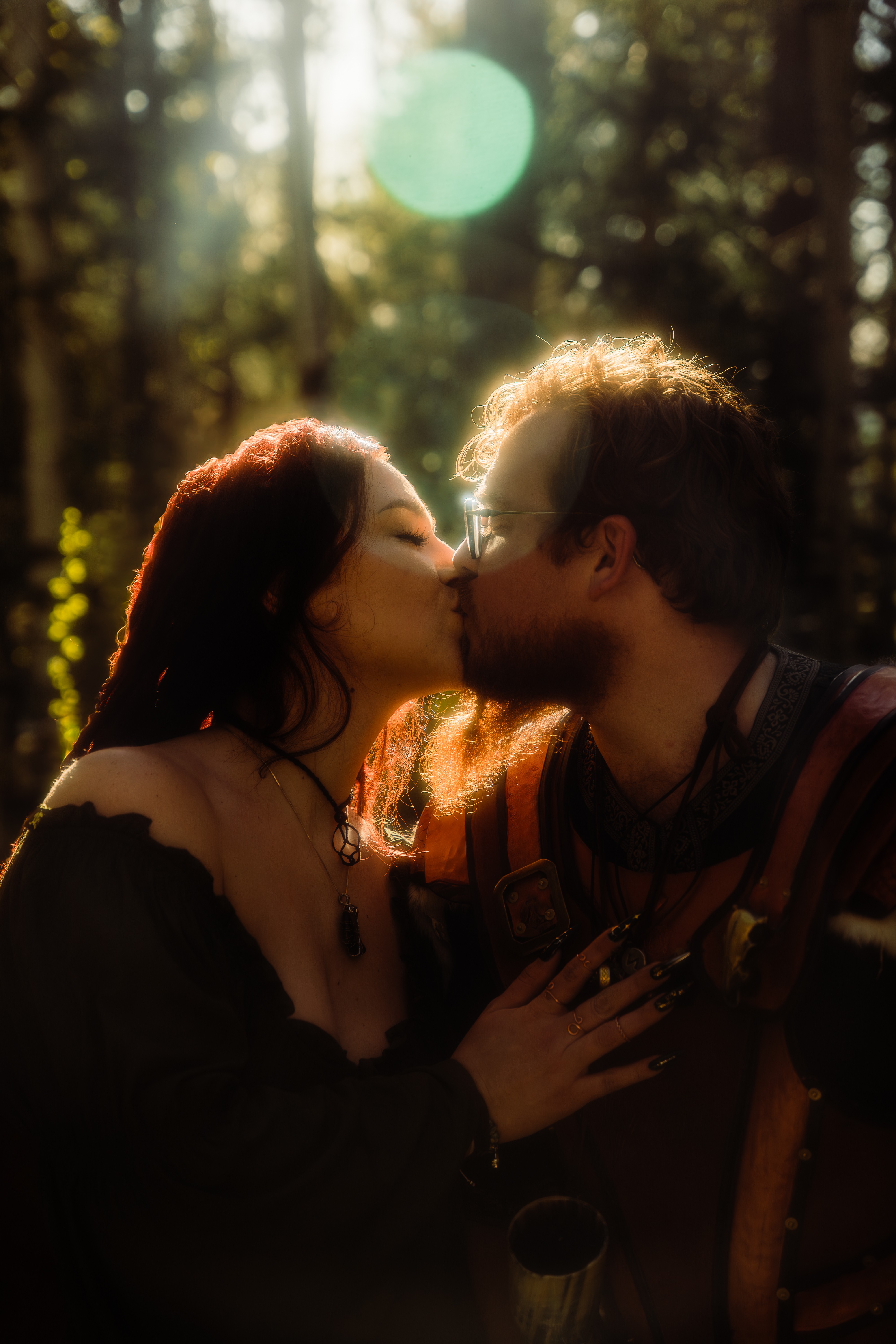 A couple kissing outdoors in a forest with sunlight filtering through trees, backlit and slightly blurred background.