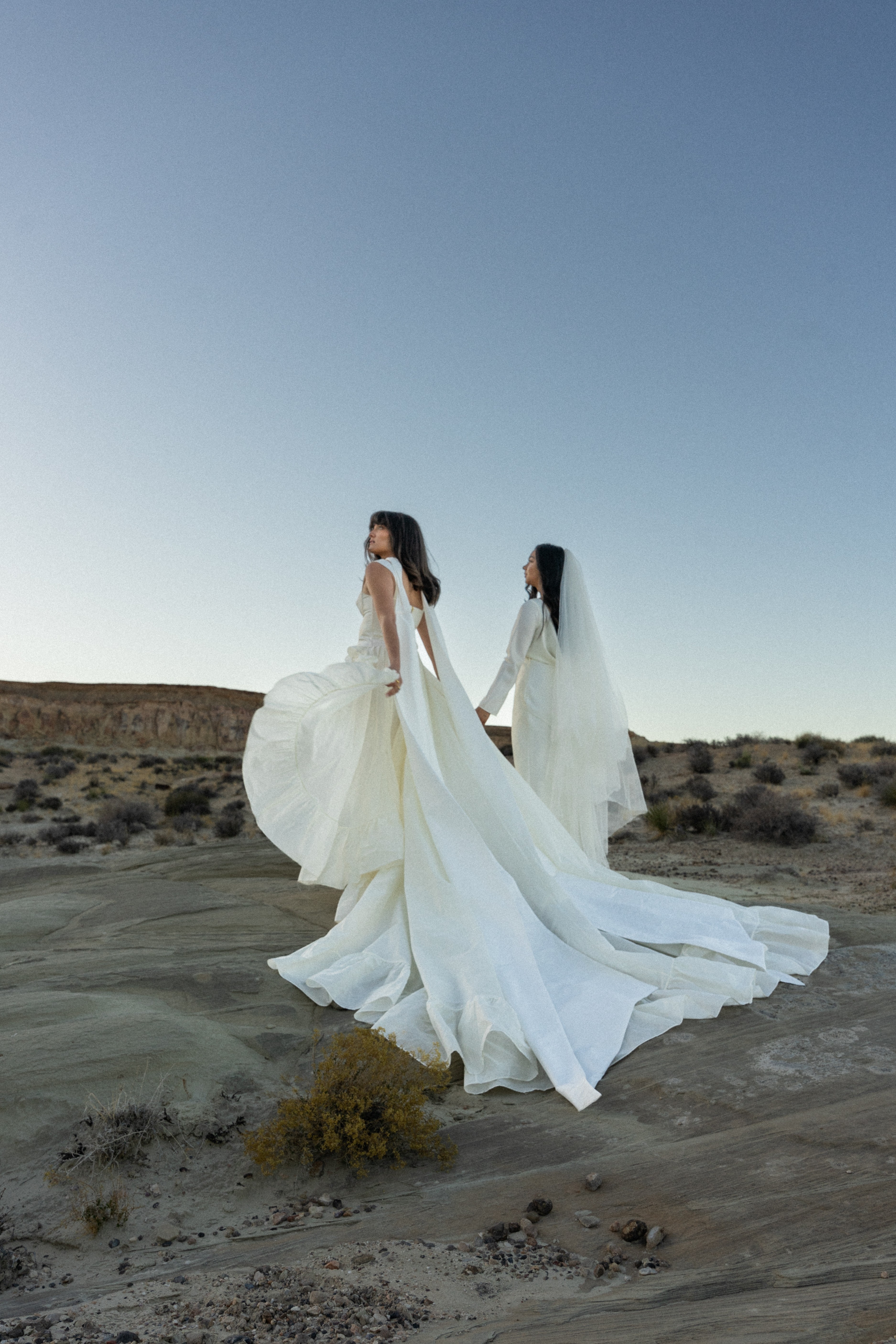 Two women in white dresses walk in a desert landscape with a clear sky, one with a long train flowing behind.