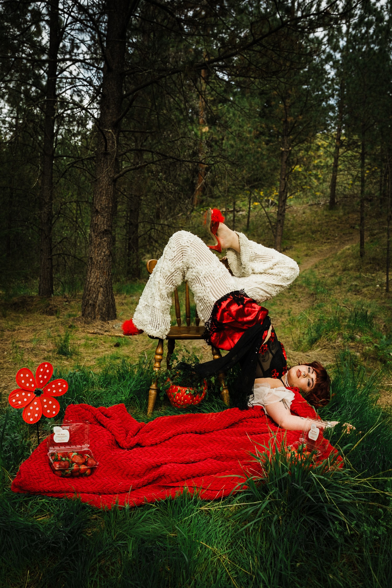 Person lying on grass with a red blanket, surrounded by trees, holding a red object, with a backpack nearby.