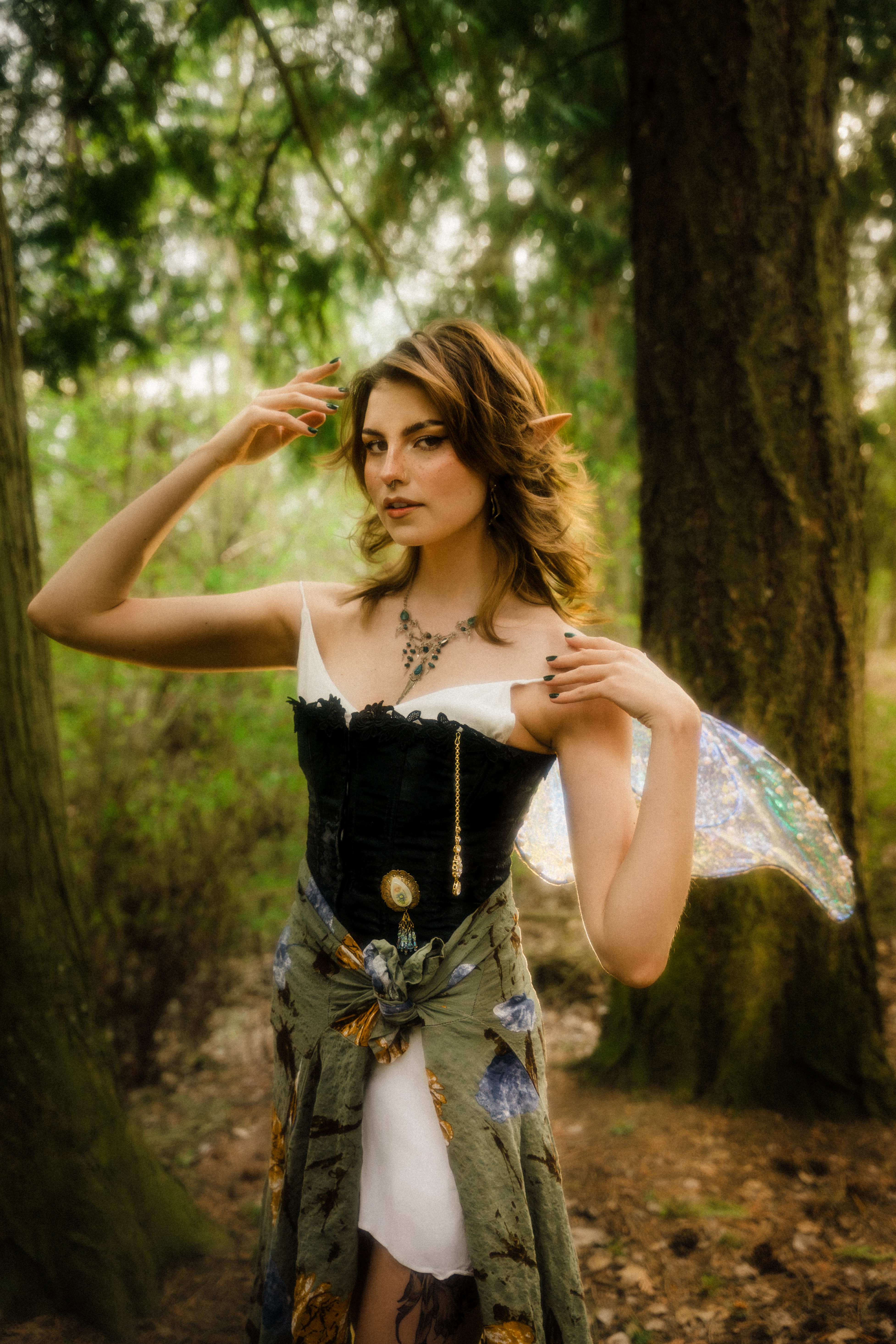 Young woman with wavy hair in a forest, wearing a black top, white skirt, and a patterned shawl, holding a necklace.