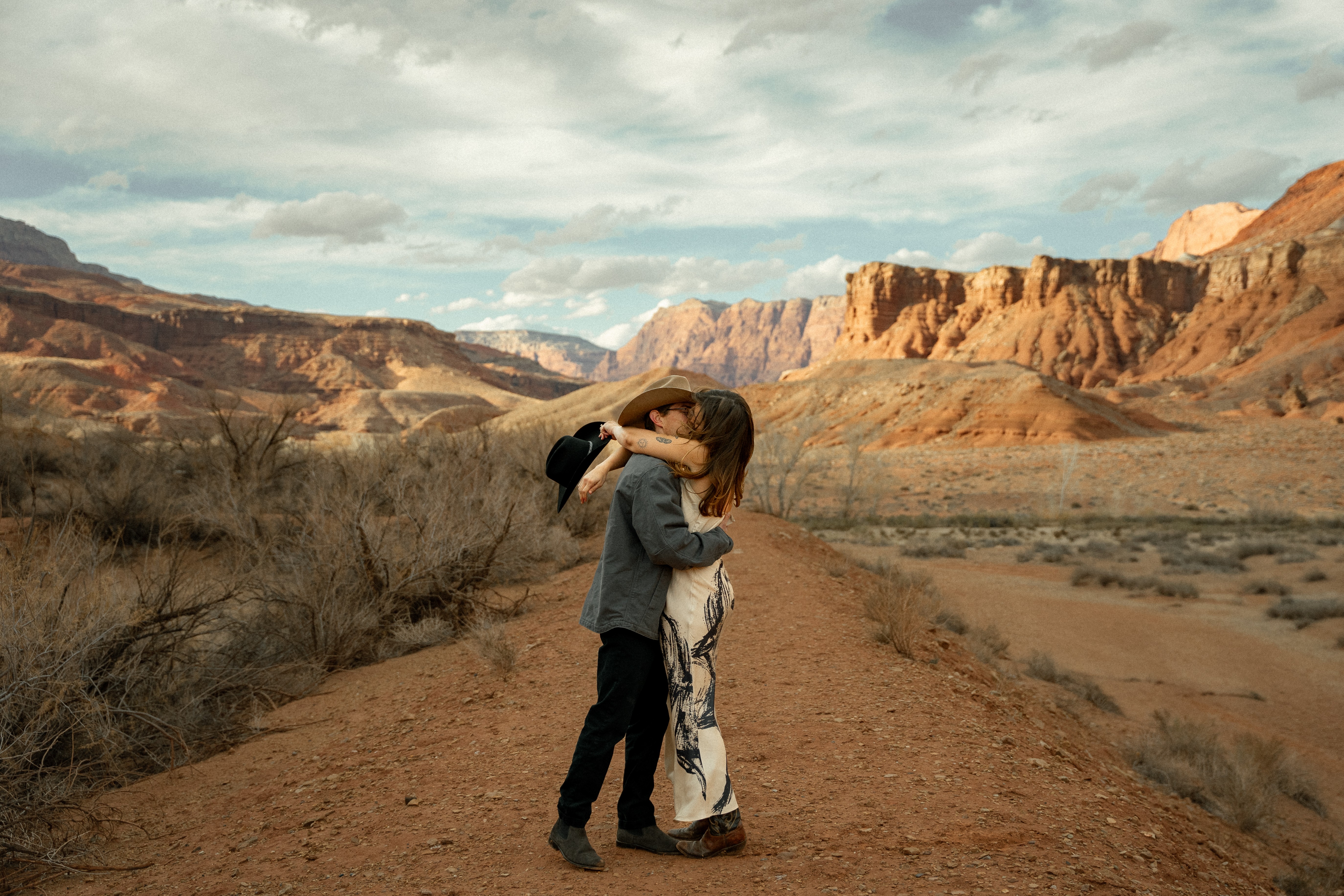 Two people hugging on a dirt path in a desert landscape with red rock formations and cloudy sky.