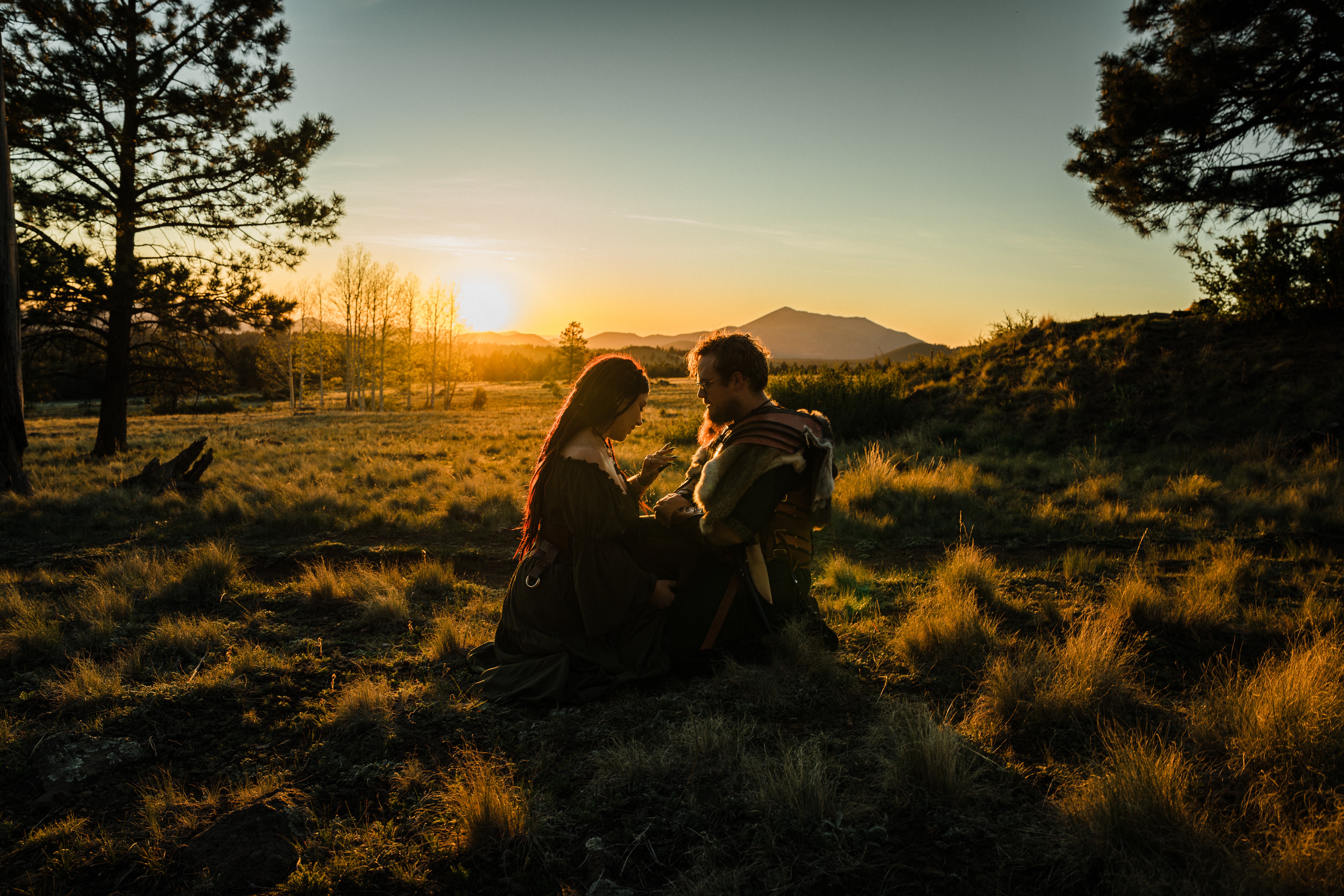 Two people kneeling and talking in a grassy field during sunset, with trees and mountains in the background.