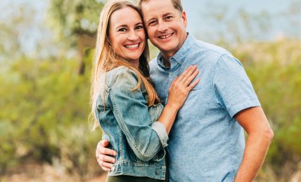 A smiling couple stands outdoors with mountains and trees in the background, embracing each other.