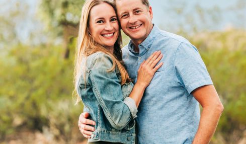 A smiling couple stands outdoors with mountains and trees in the background, embracing each other.