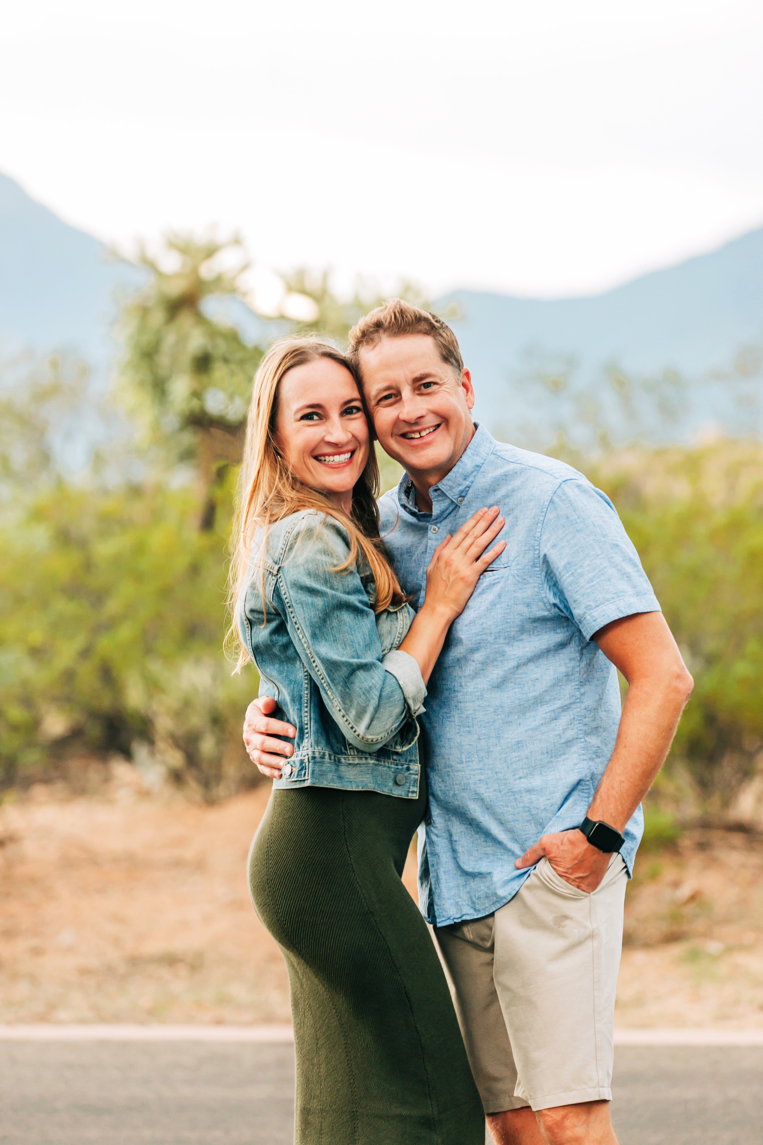 A smiling couple stands outdoors with mountains and trees in the background, embracing each other.