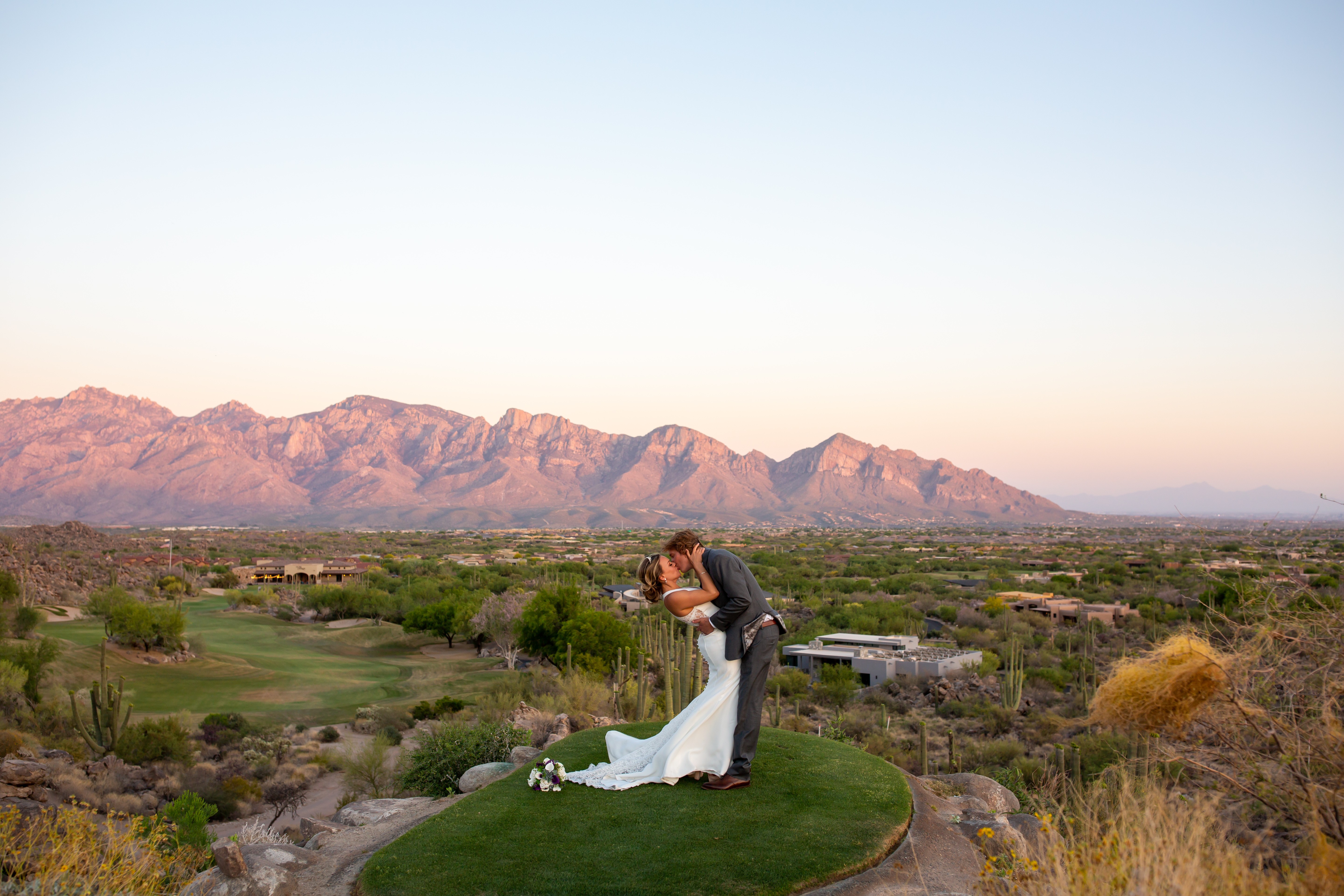 A couple in wedding attire standing on a grassy area with mountains in the background during sunset.