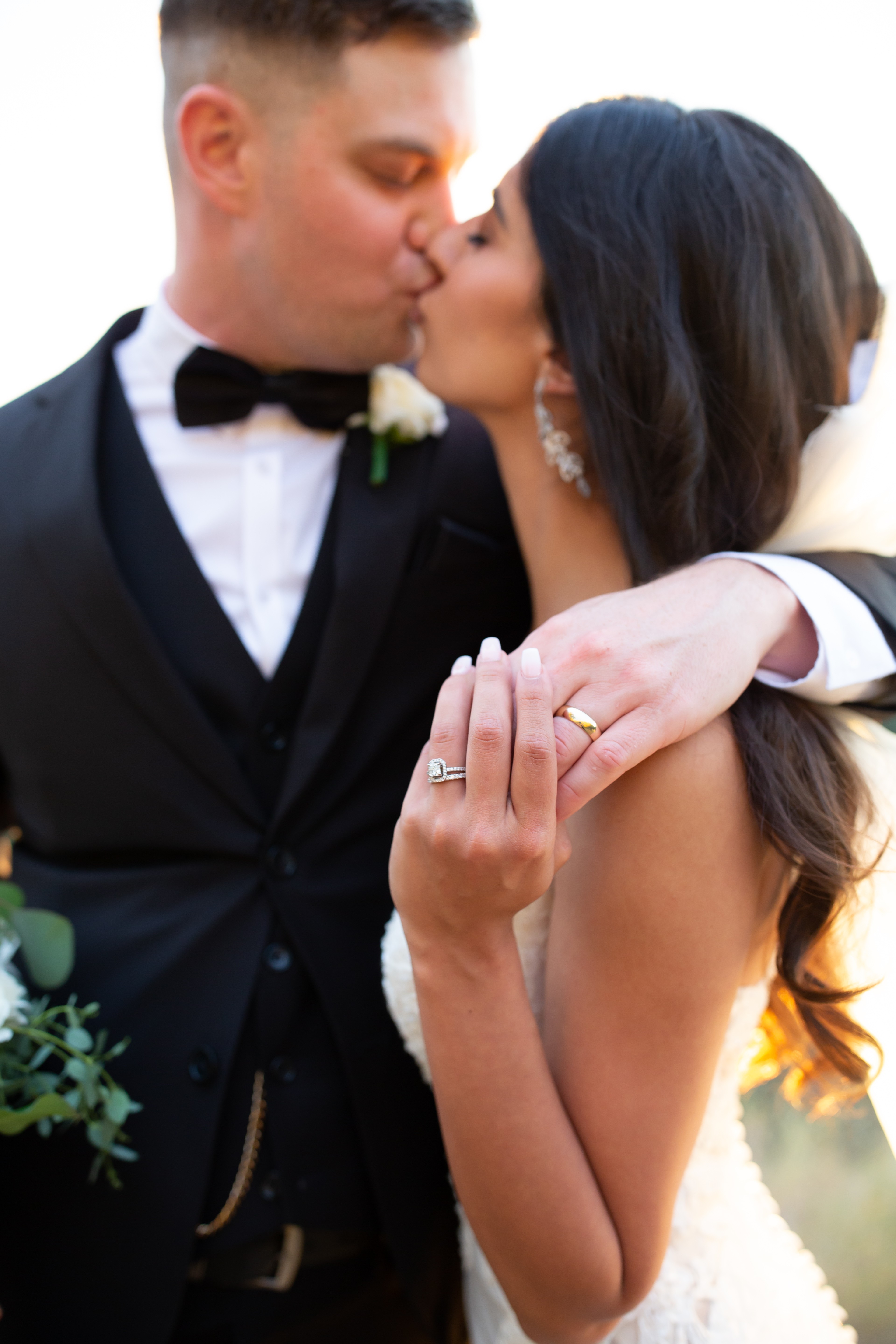 A bride and groom kiss, the groom in a tuxedo and the bride with long dark hair, holding her hand with a wedding ring.