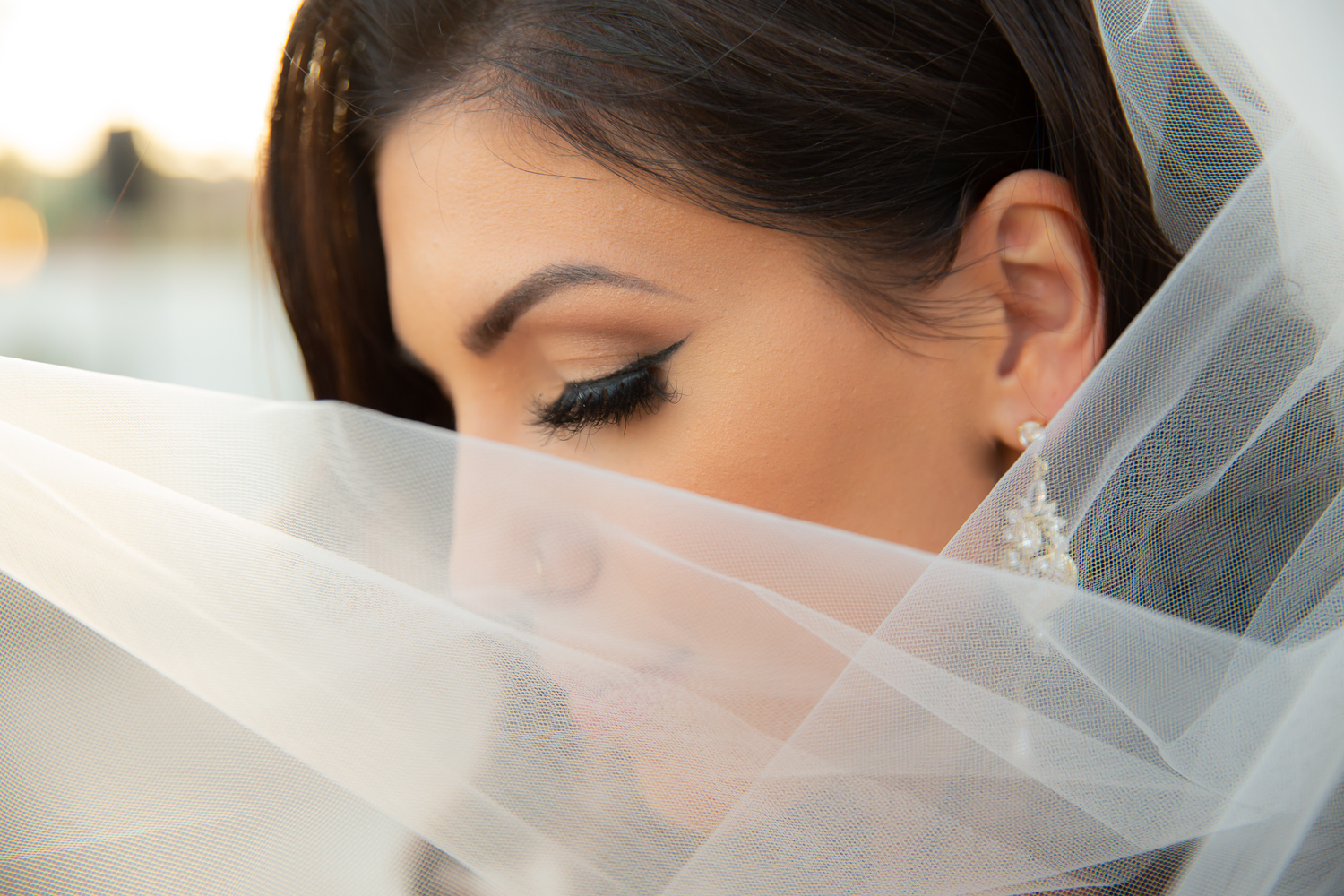 Close-up of a woman with makeup and earrings, partially covered by sheer fabric, outdoors during sunset.
