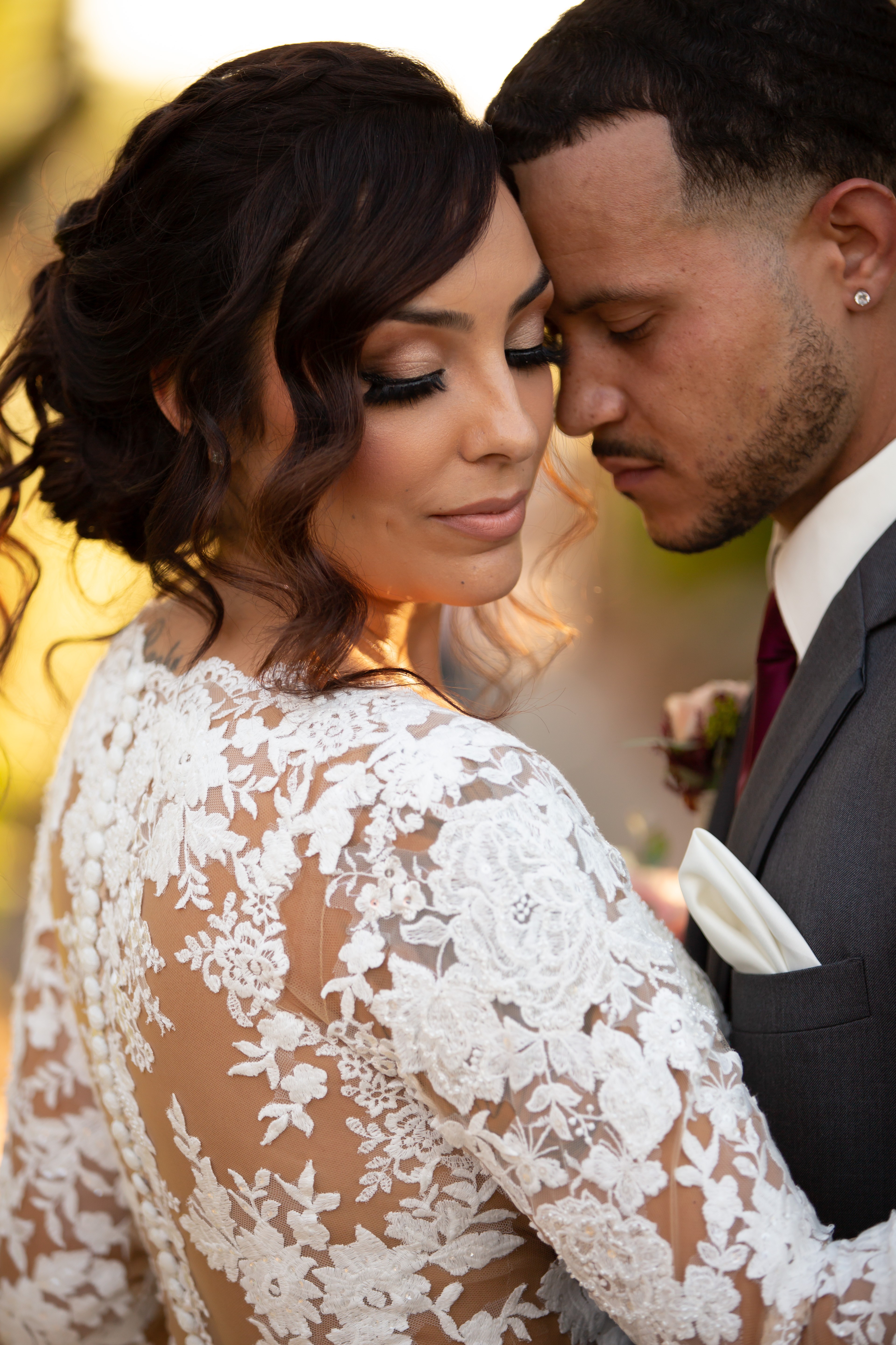 Close-up of a bride and groom with foreheads touching, eyes closed, outdoors, bride in lace dress, groom in suit.
