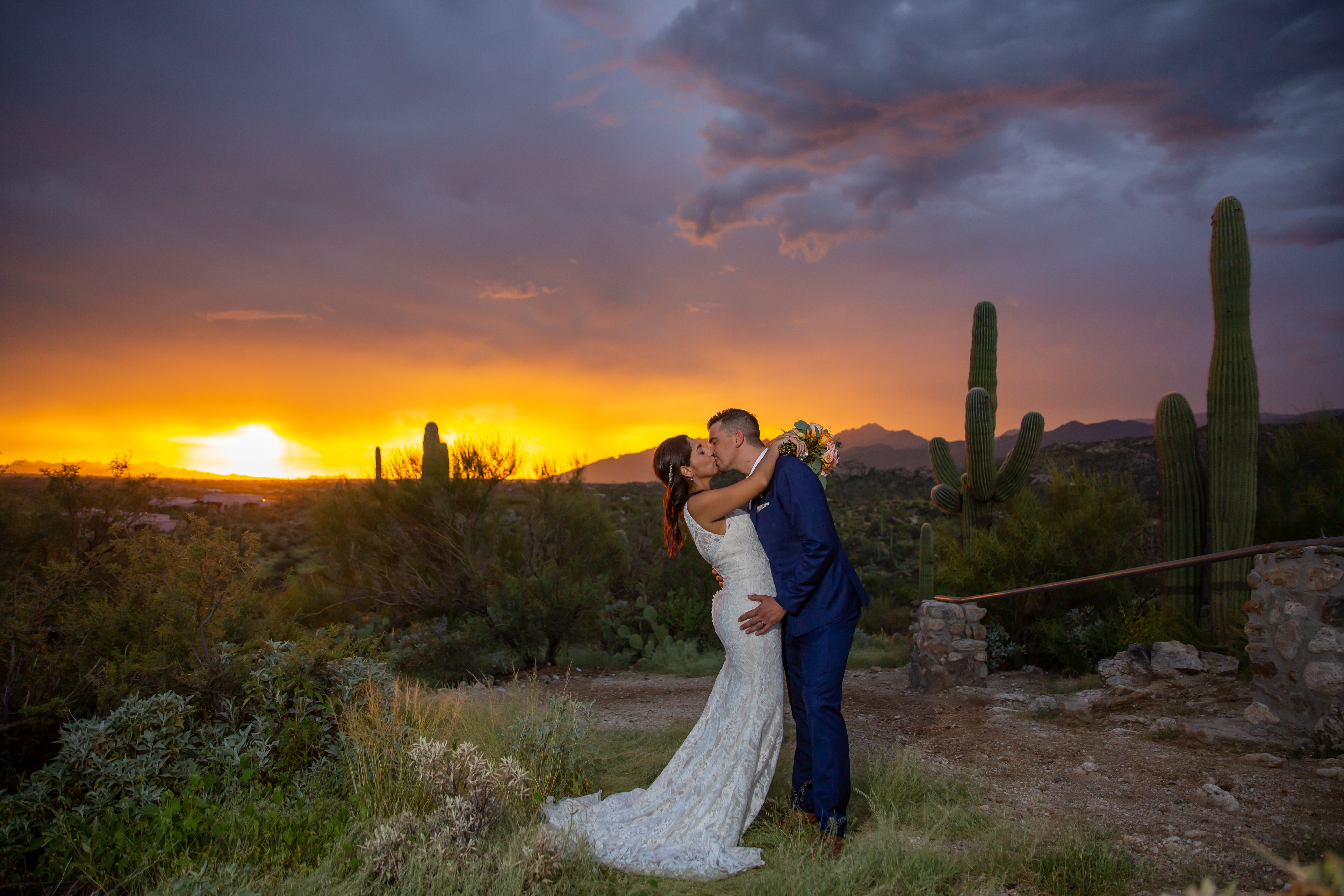 A couple in wedding attire kissing outdoors at sunset with cacti and desert landscape in background.