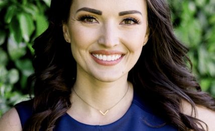 Young woman with long dark hair smiling, wearing a blue top, outdoors with green foliage background.