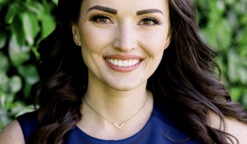 Young woman with long dark hair smiling, wearing a blue top, outdoors with green foliage background.