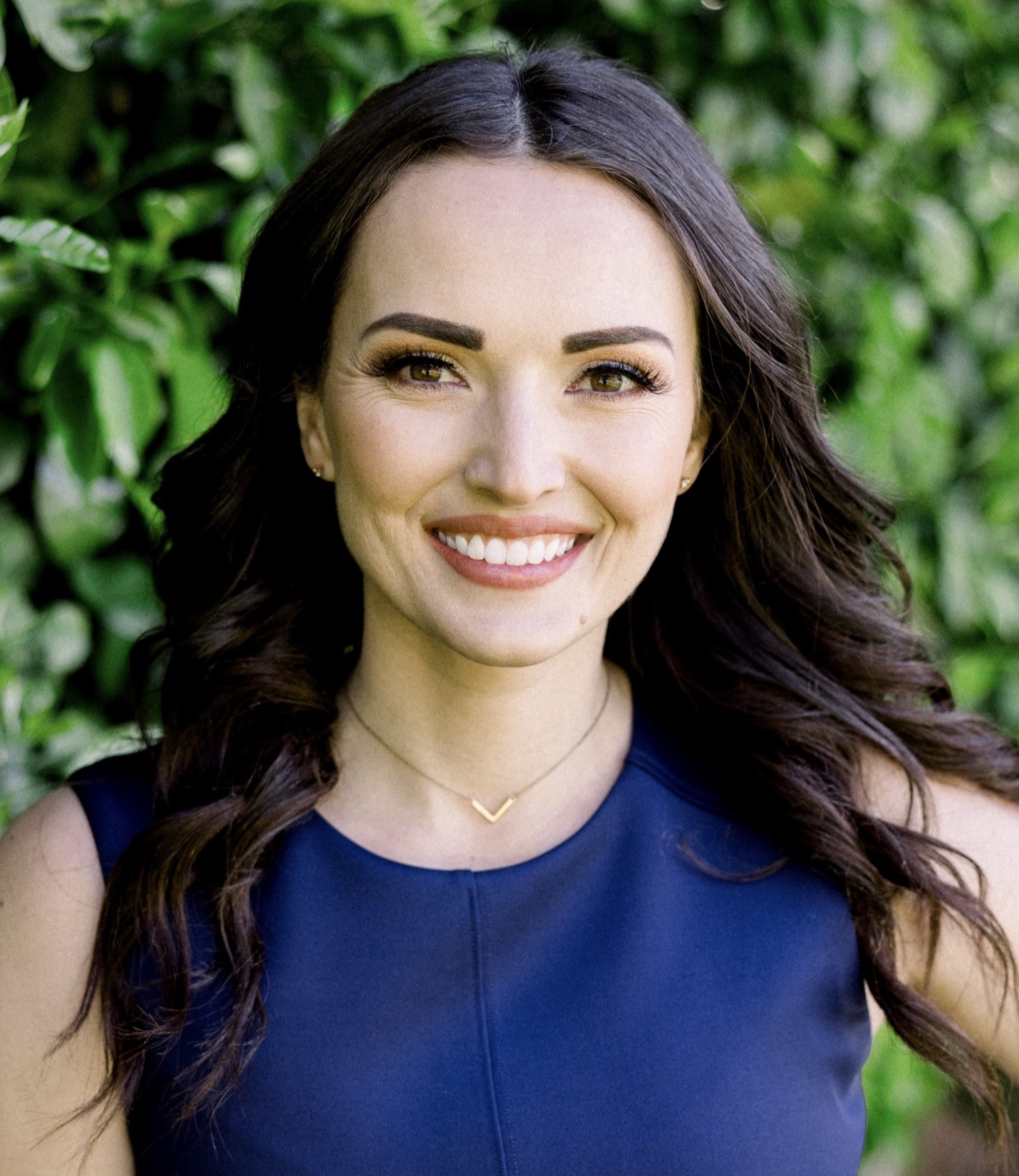 Young woman with long dark hair smiling, wearing a blue top, outdoors with green foliage background.