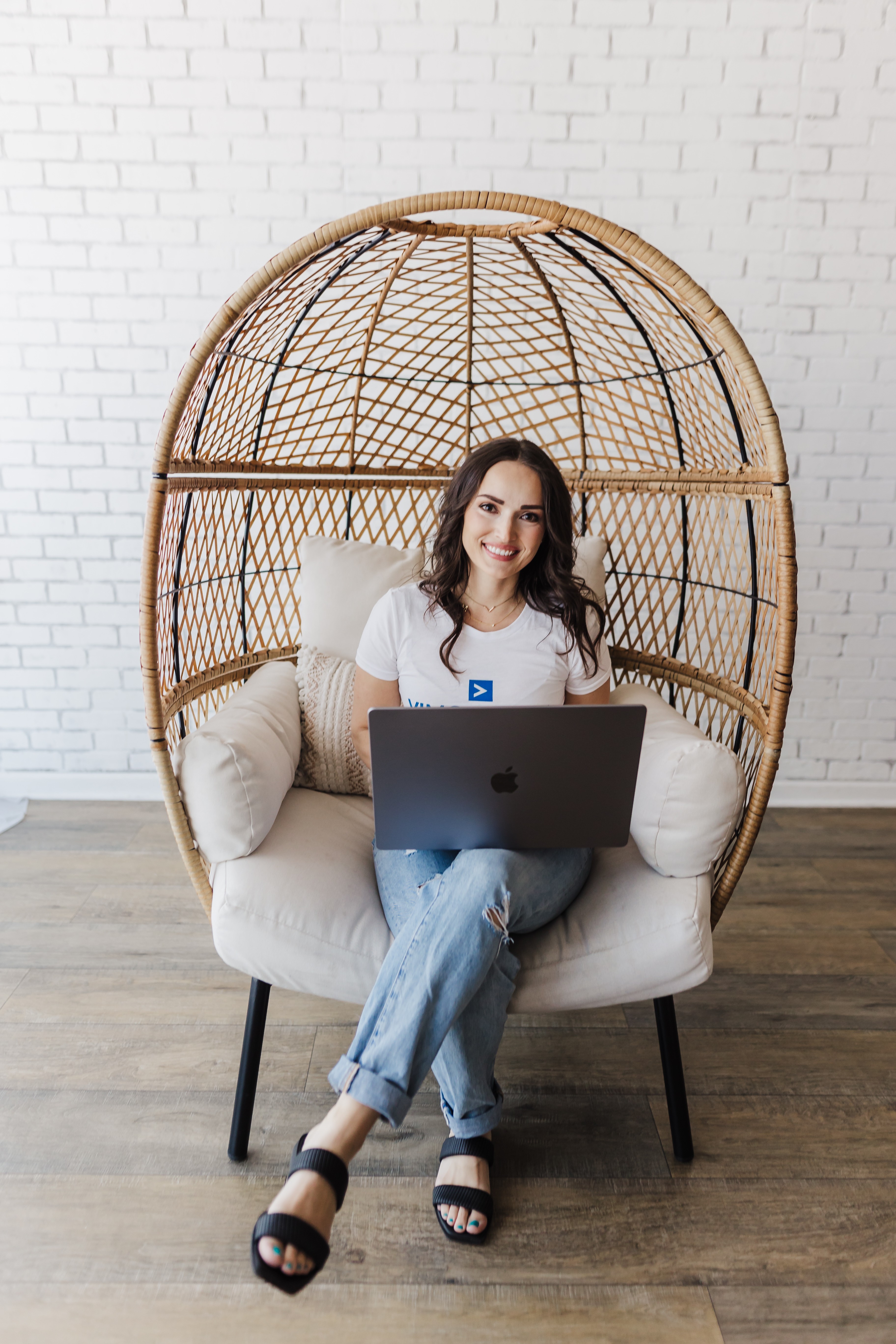 Woman sitting on a cushioned chair with a laptop, inside a wicker dome-shaped structure, against a white brick wall.