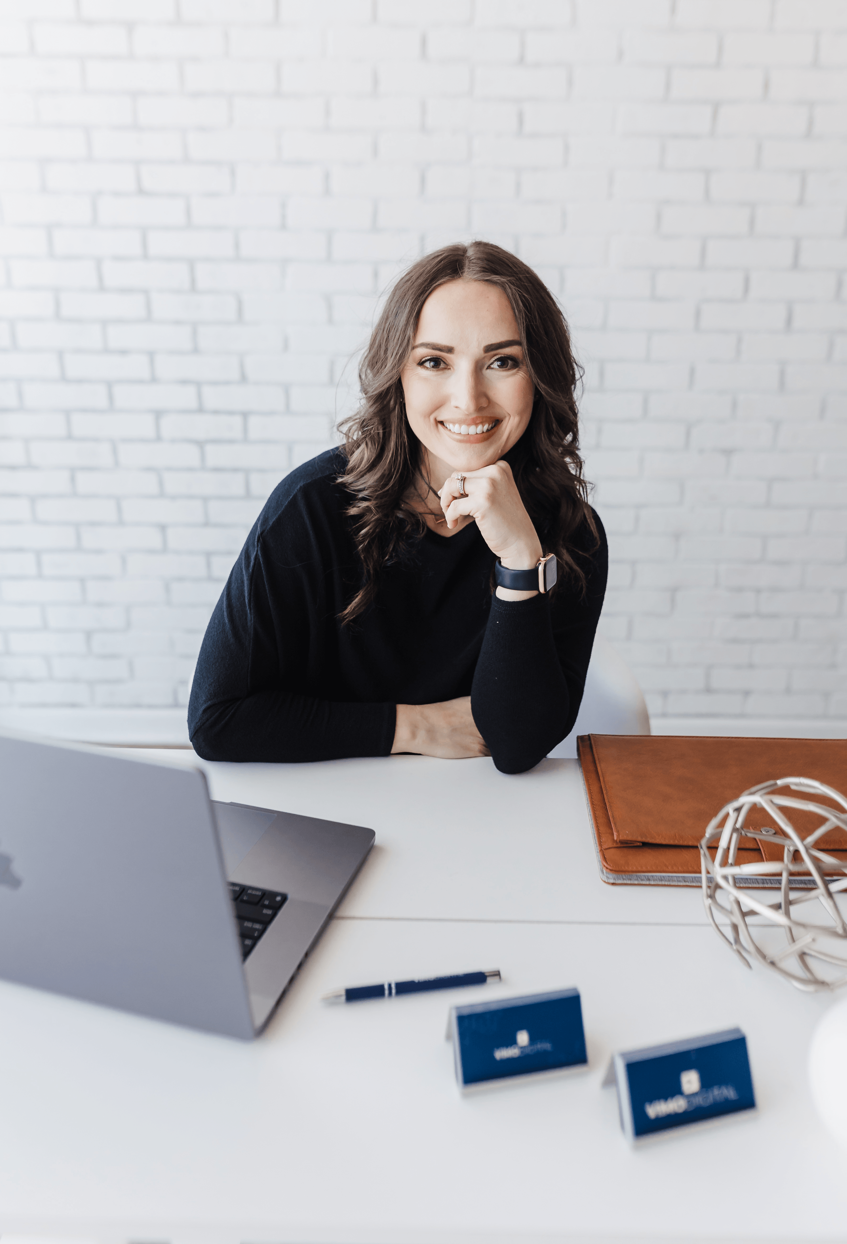 Smiling woman sitting at desk with laptop, notebook, and decorative object, against a white brick wall background.
