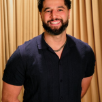 Smiling man with curly hair and beard, wearing a dark shirt, standing in front of a gold curtain.