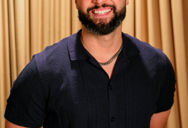 Smiling man with curly hair and beard, wearing a dark shirt, standing in front of a gold curtain.