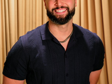 Smiling man with curly hair and beard, wearing a dark shirt, standing in front of a gold curtain.