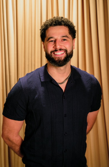 Smiling man with curly hair and beard, wearing a dark shirt, standing in front of a gold curtain.