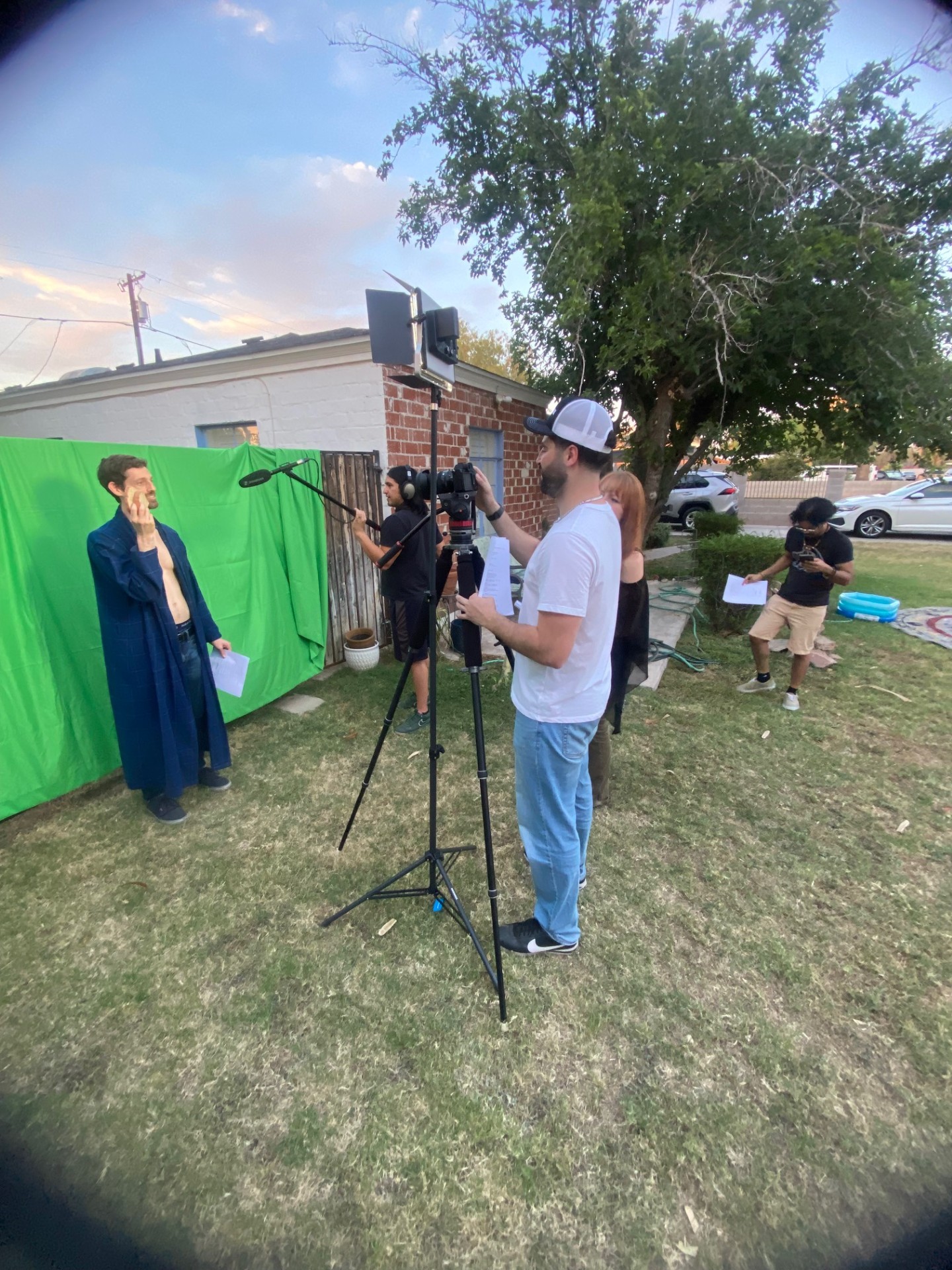 Person in white shirt and cap filming a woman speaking in front of a green screen outdoors, with crew members and equipment nearby.