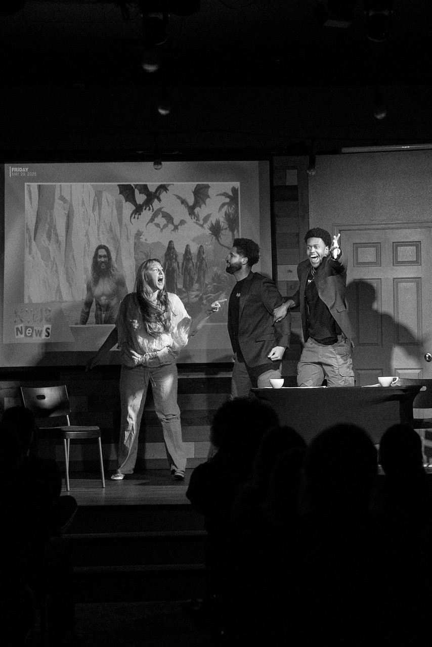 Three people stand on stage in front of a screen displaying a forest scene with bats, in a dark room with an audience.