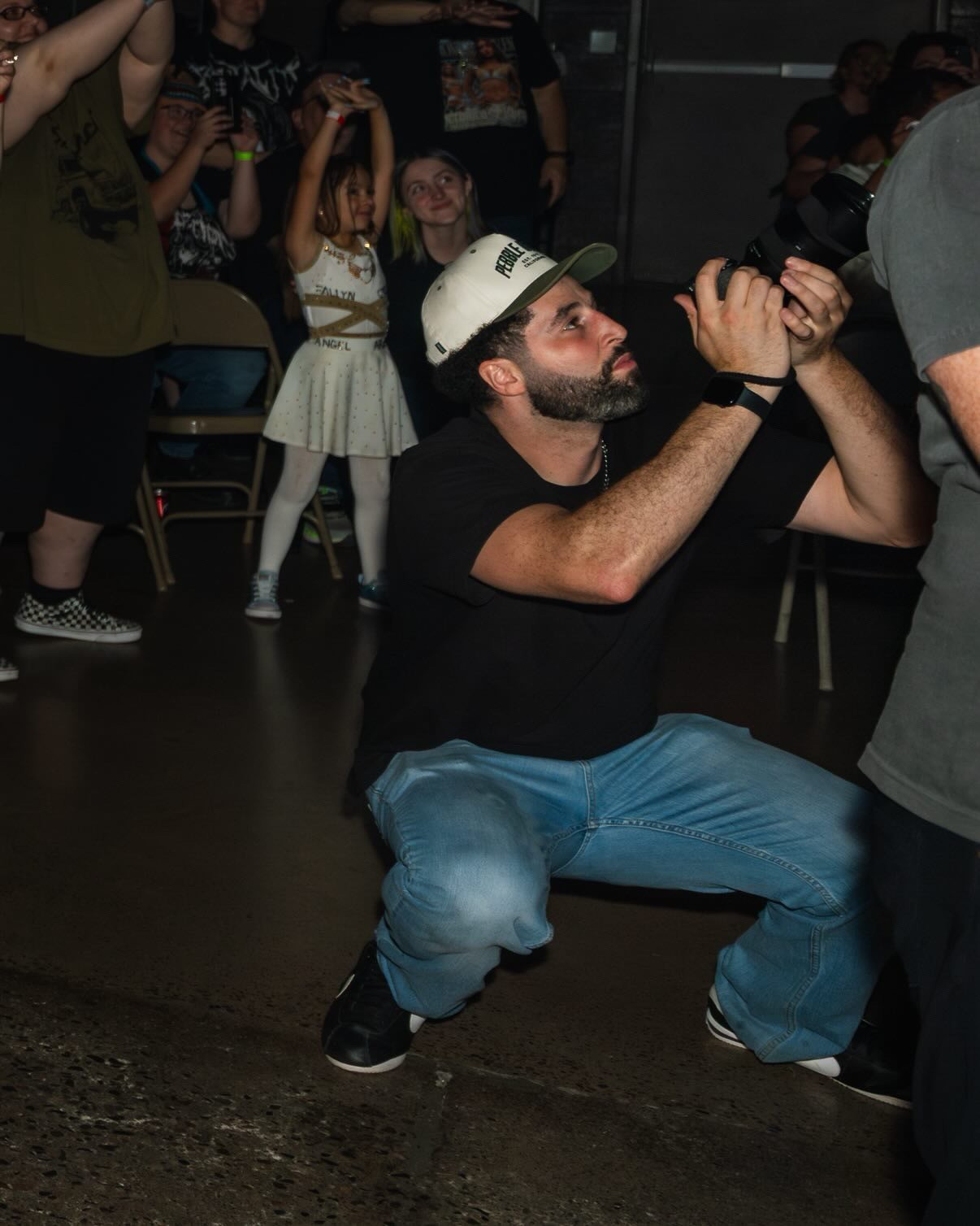 Man crouching and holding a microphone, with children and people in the background at an indoor event.