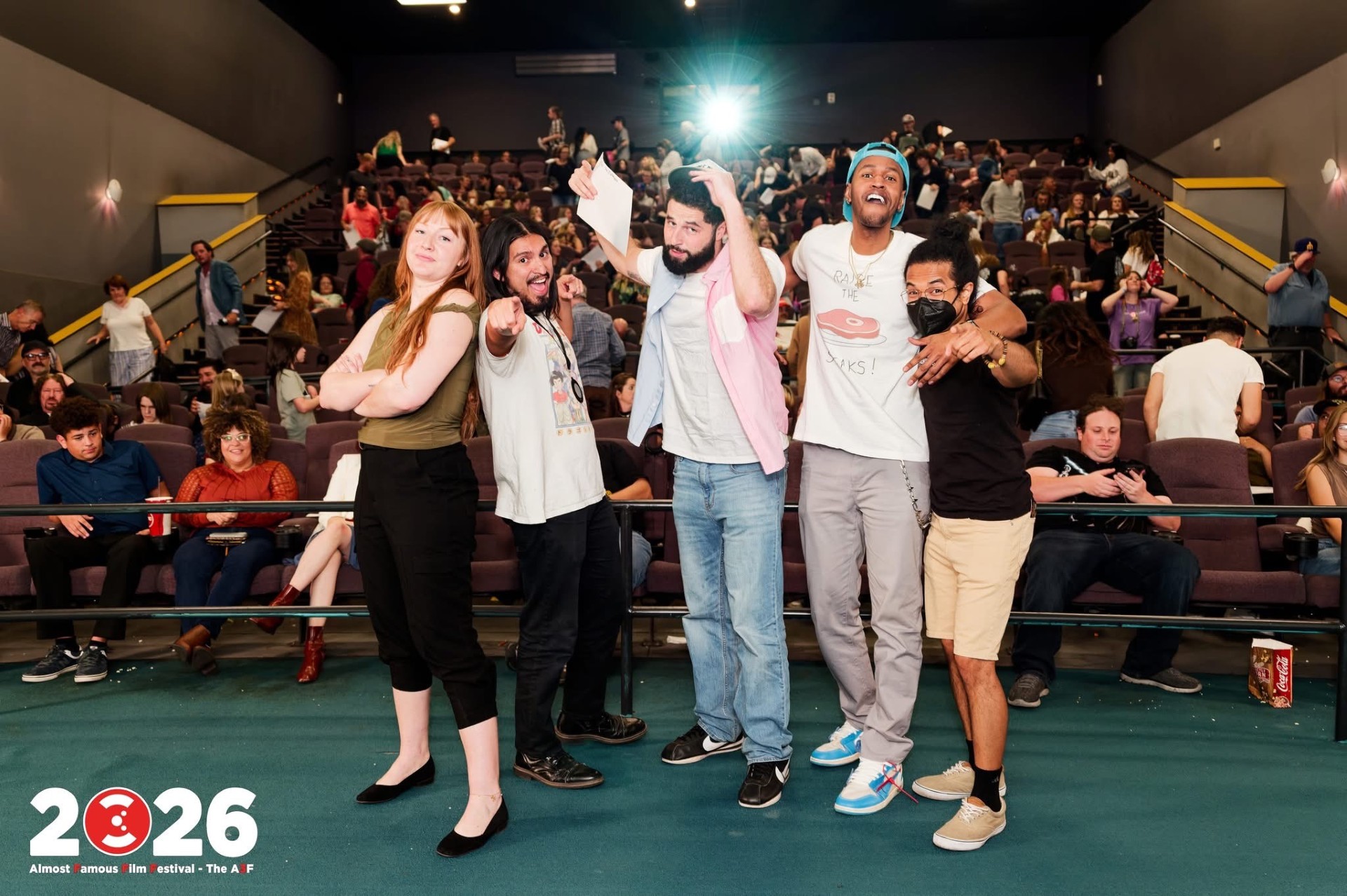 Group of five people standing on stage in front of an audience, some with arms around each other, in a theater setting.