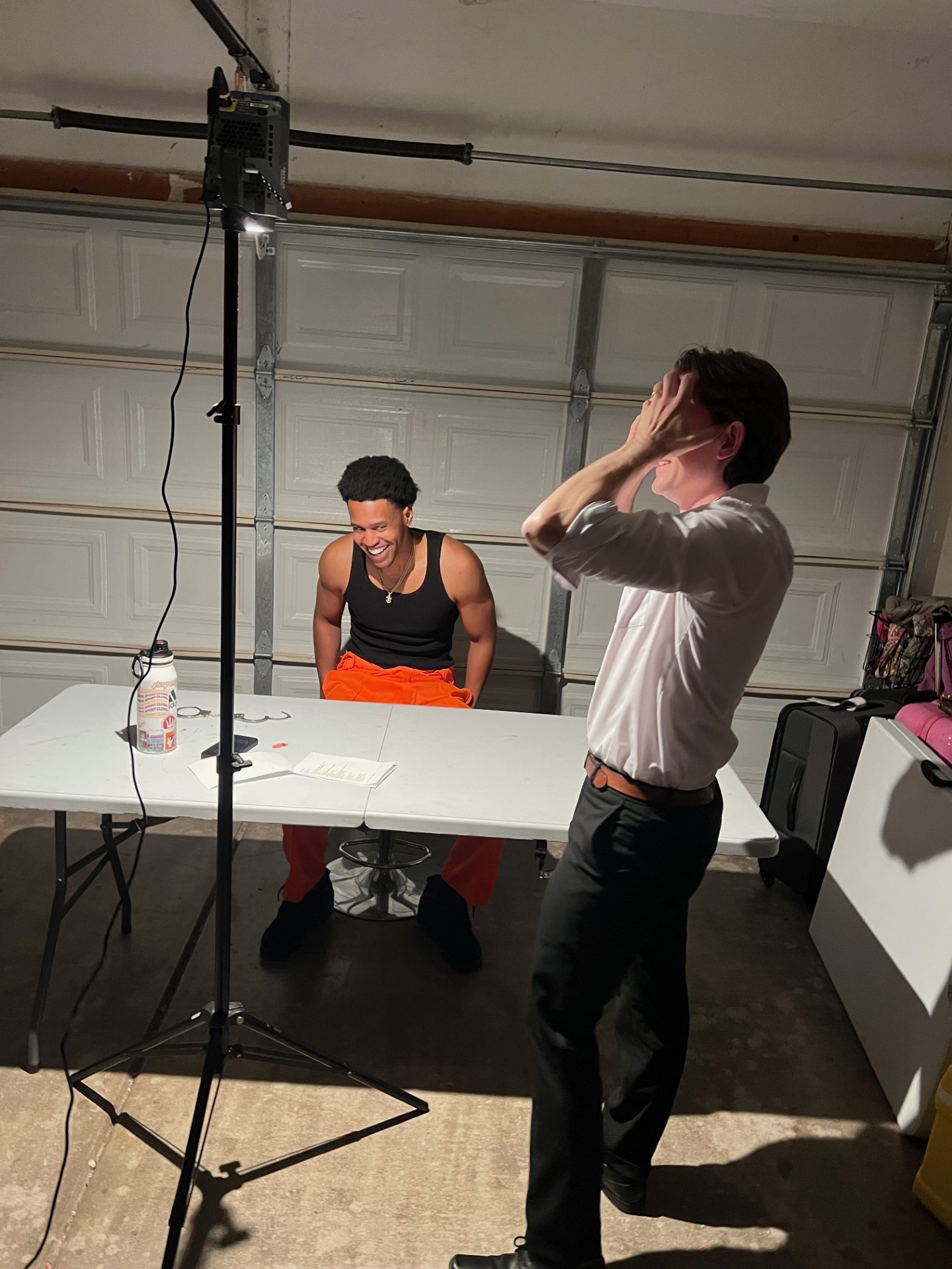 Two people in a garage, one sitting at a table and the other standing, adjusting hair or glasses, with studio lighting equipment nearby.