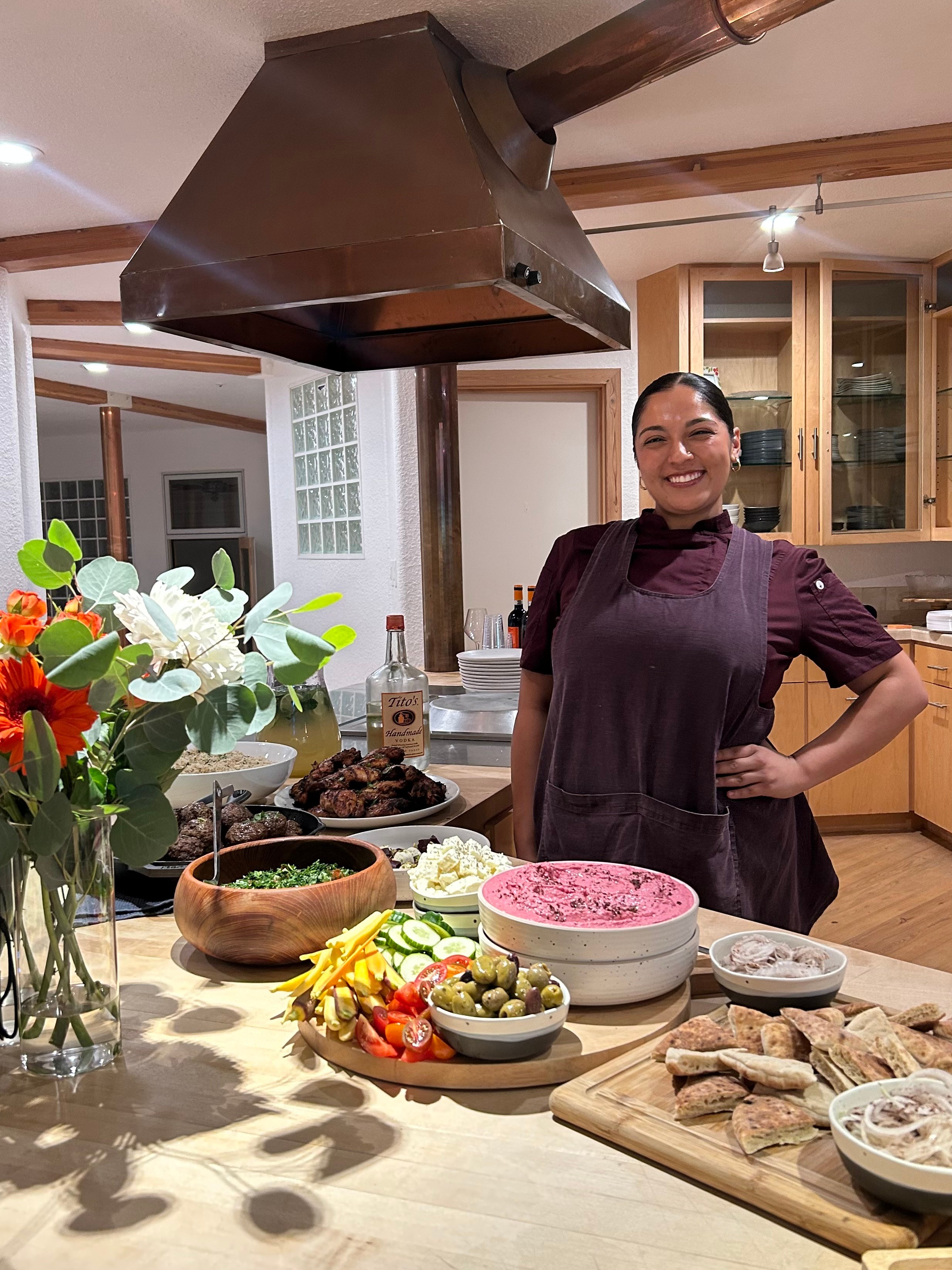 Smiling woman in a kitchen with a table of food, flowers, and a wooden cutting board, under a large range hood.