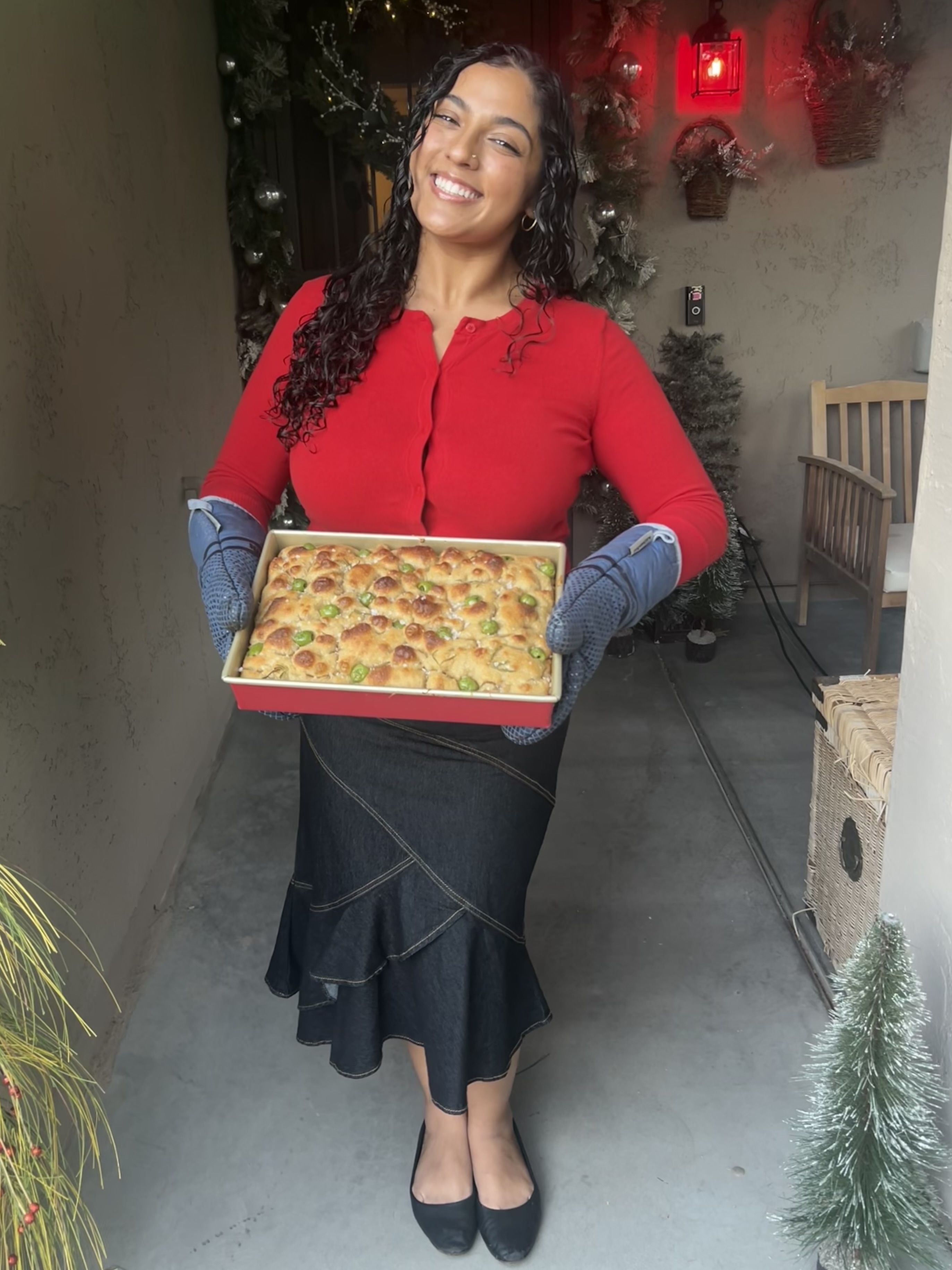 Woman in red sweater holding a pizza box, smiling, standing indoors with plants and furniture nearby.