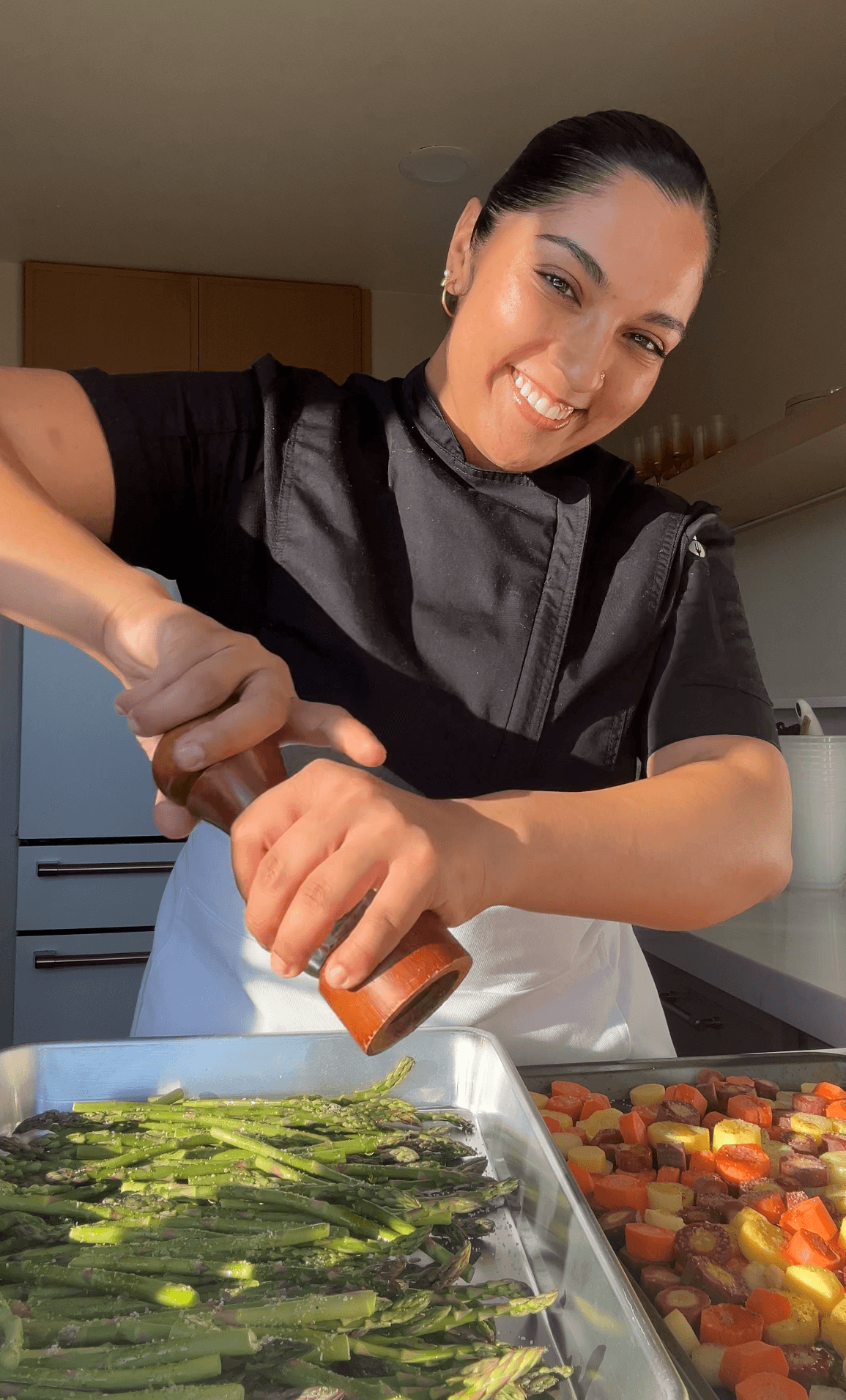 Woman smiling while seasoning vegetables in a kitchen, with a tray of roasted vegetables in front.