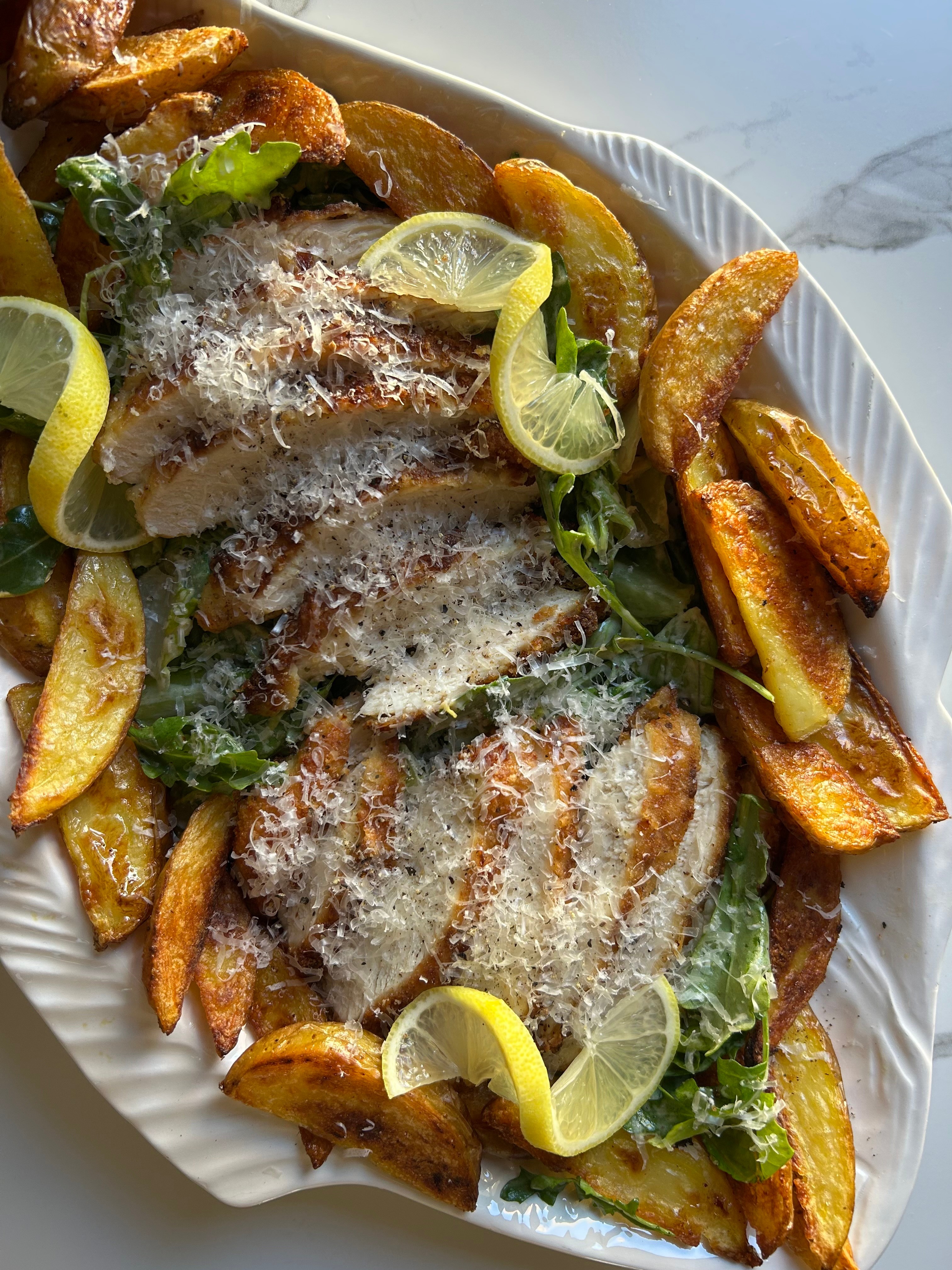 Grilled fish with lemon slices, served with potato wedges and greens on a white plate.