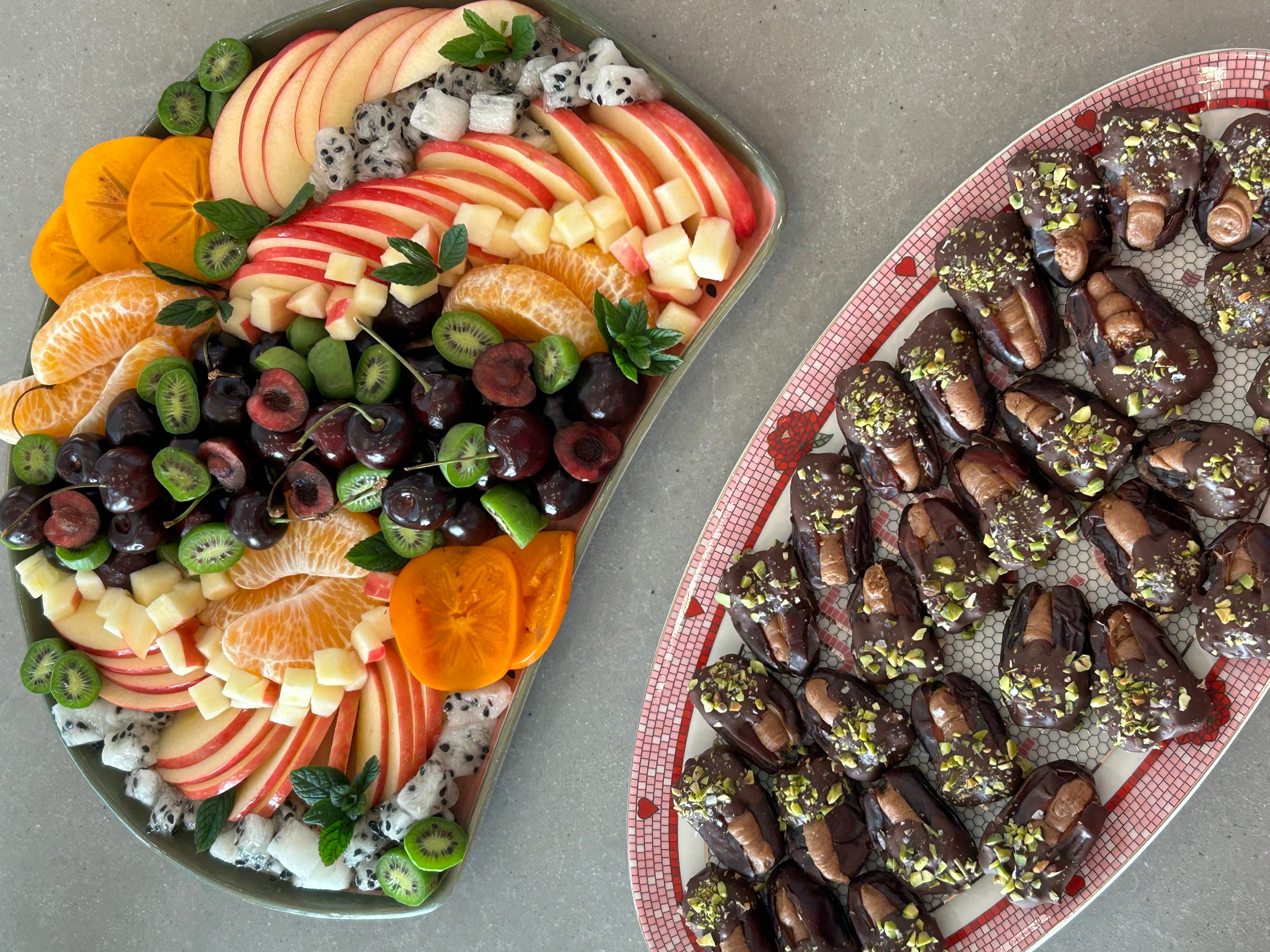 Assorted fruit platter with sliced apples, oranges, grapes, and melon, alongside a plate of chocolate-covered treats with nuts and sprinkles.