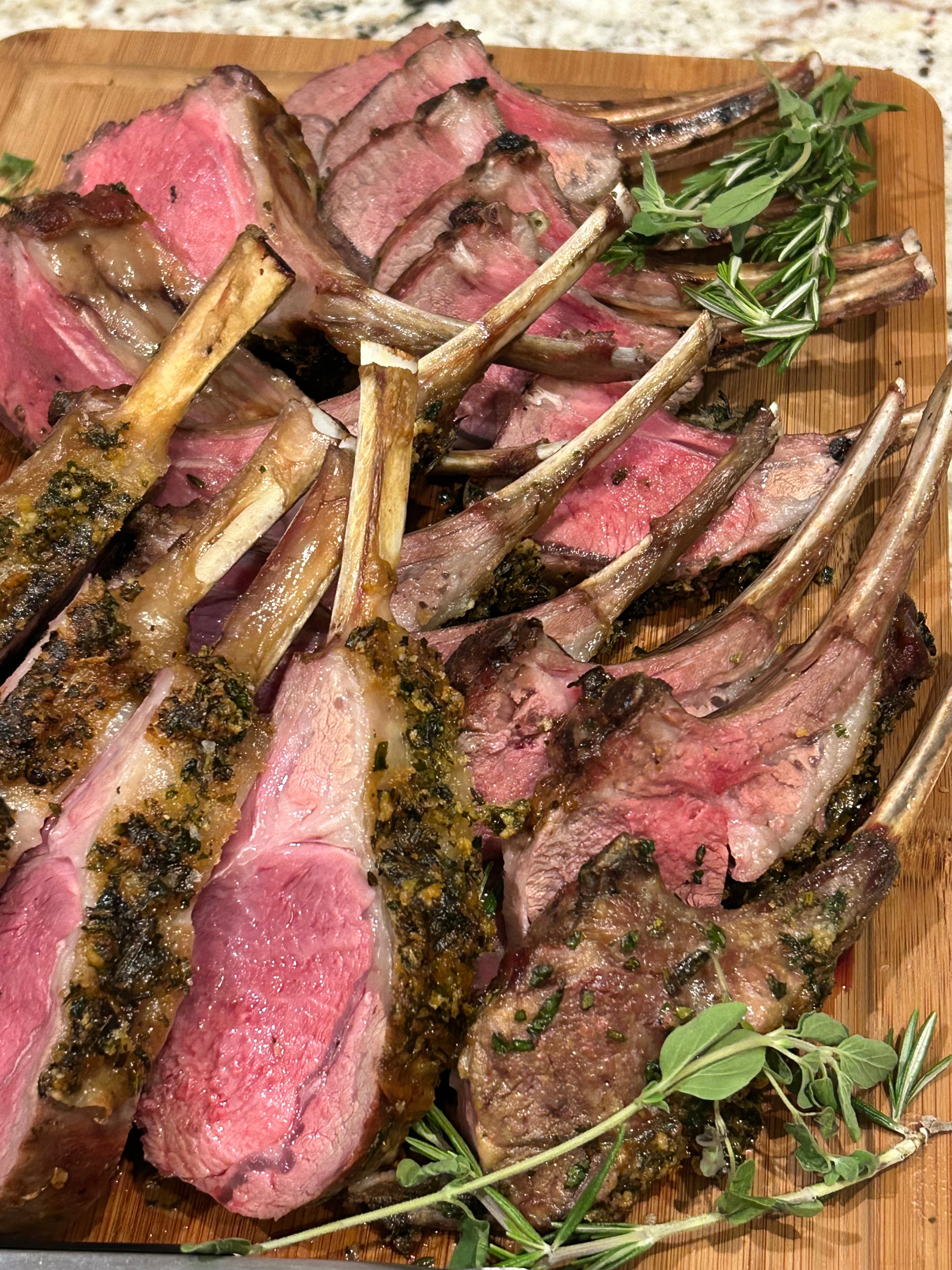 Slices of cooked lamb with herbs on a wooden platter, garnished with sprigs of rosemary and other herbs.