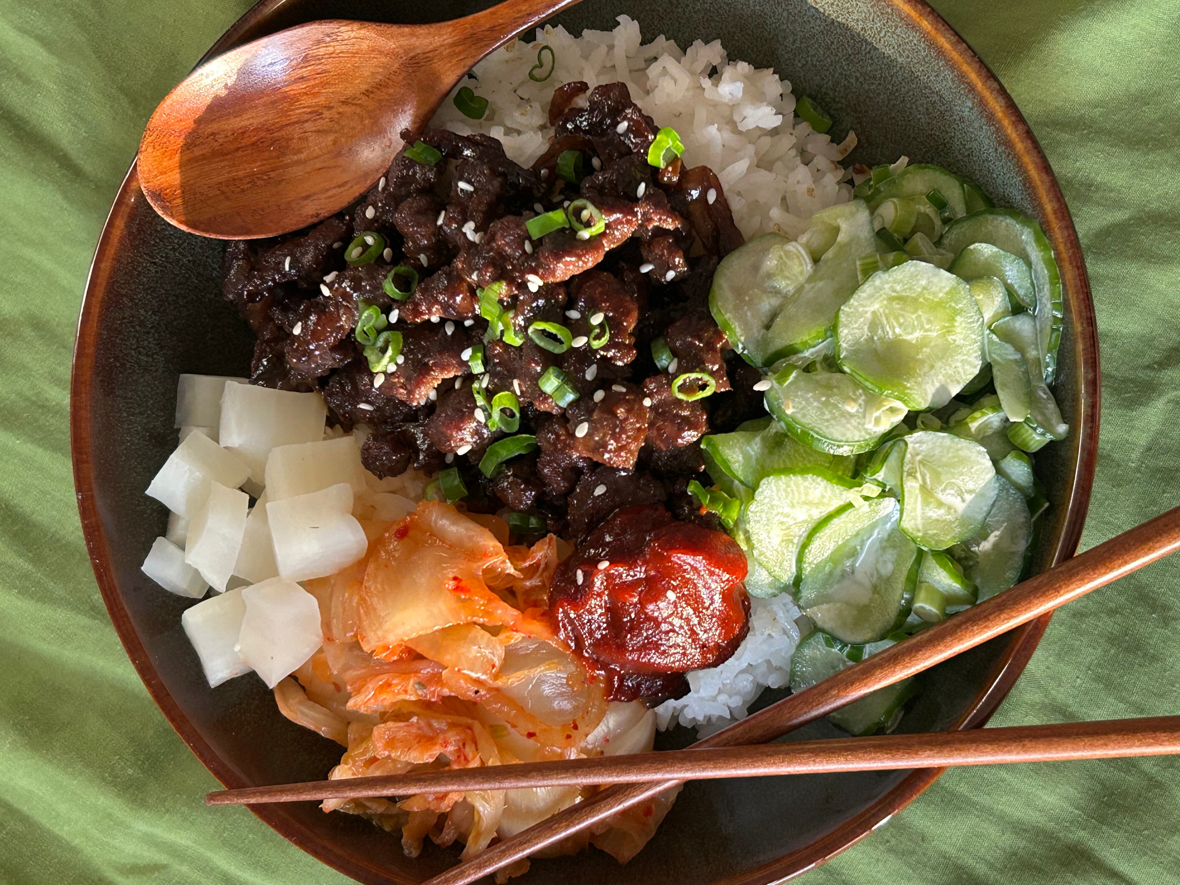 Bowl with rice, cooked beef, cucumber slices, kimchi, and a spoon and chopsticks, on a green cloth.